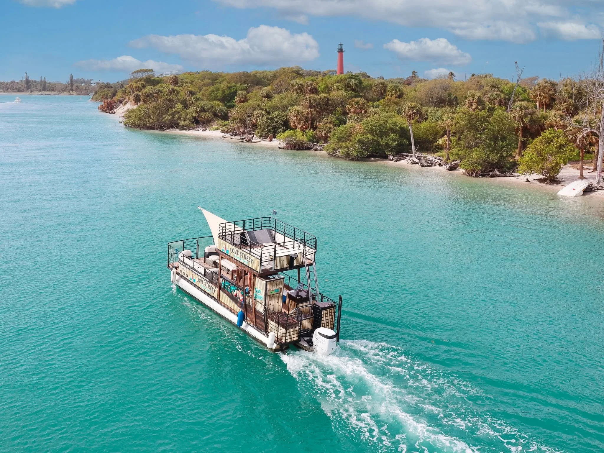 A boat with a sign reading 'Love Street' cruises on turquoise water near a green, tree-lined shoreline with a lighthouse in the distance.