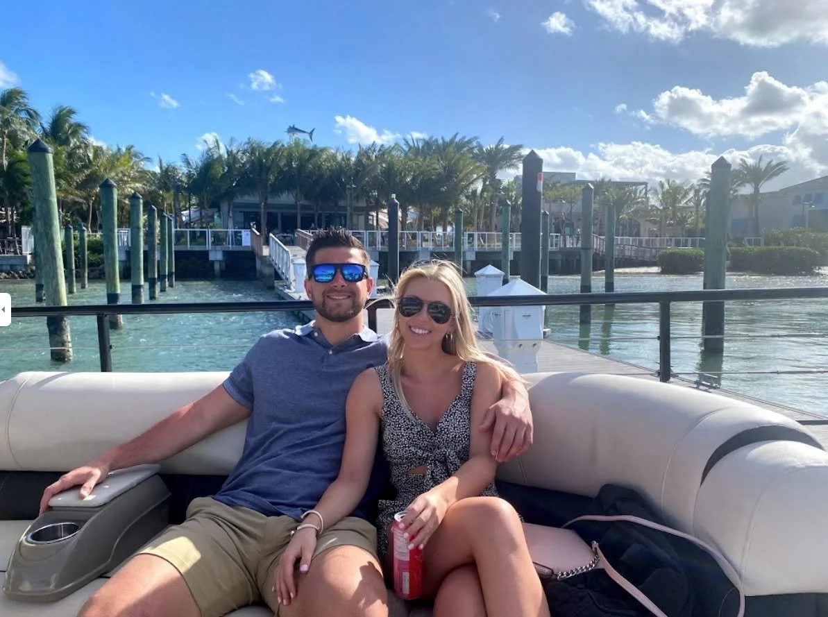 A man and woman sitting together on a boat deck with water and palm trees in the background, both wearing sunglasses and smiling.