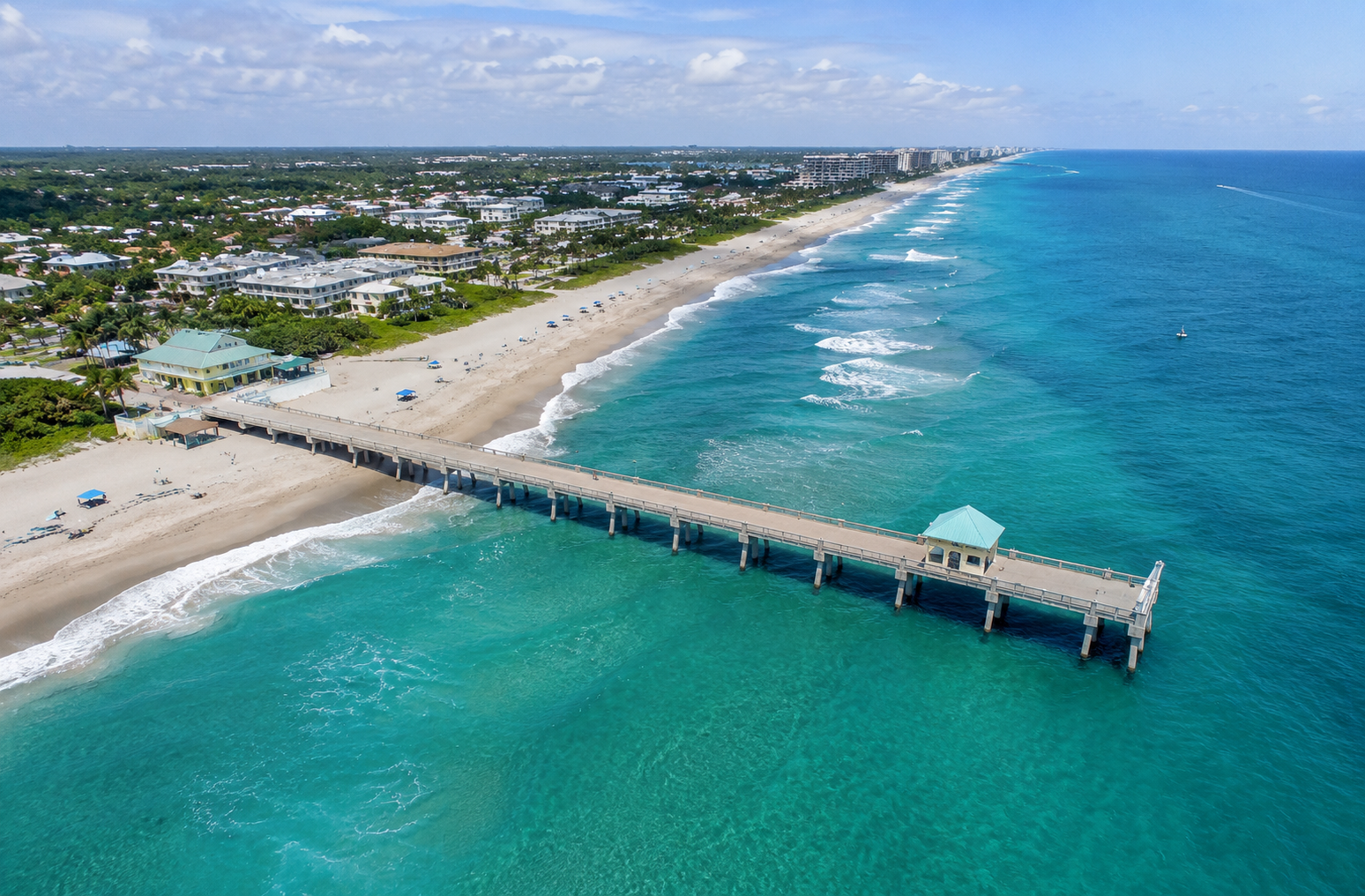 Aerial view of a long pier extending out into the ocean with a small building at the end, sandy beach, and a residential area with houses and trees along the coastline on a sunny day with clouds in the sky.