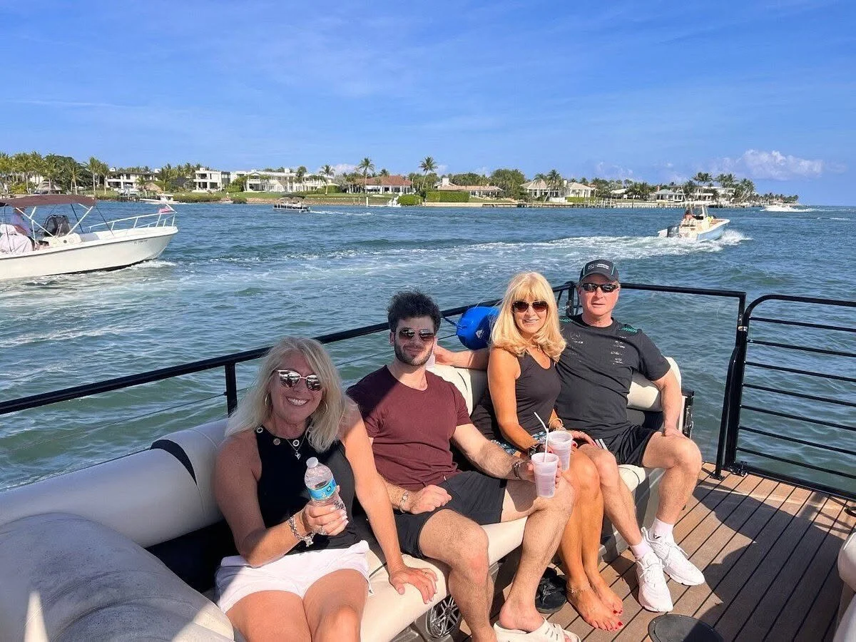 Four adults sitting on a boat enjoying a day on the water, with houses, boats, and palm trees in the background.