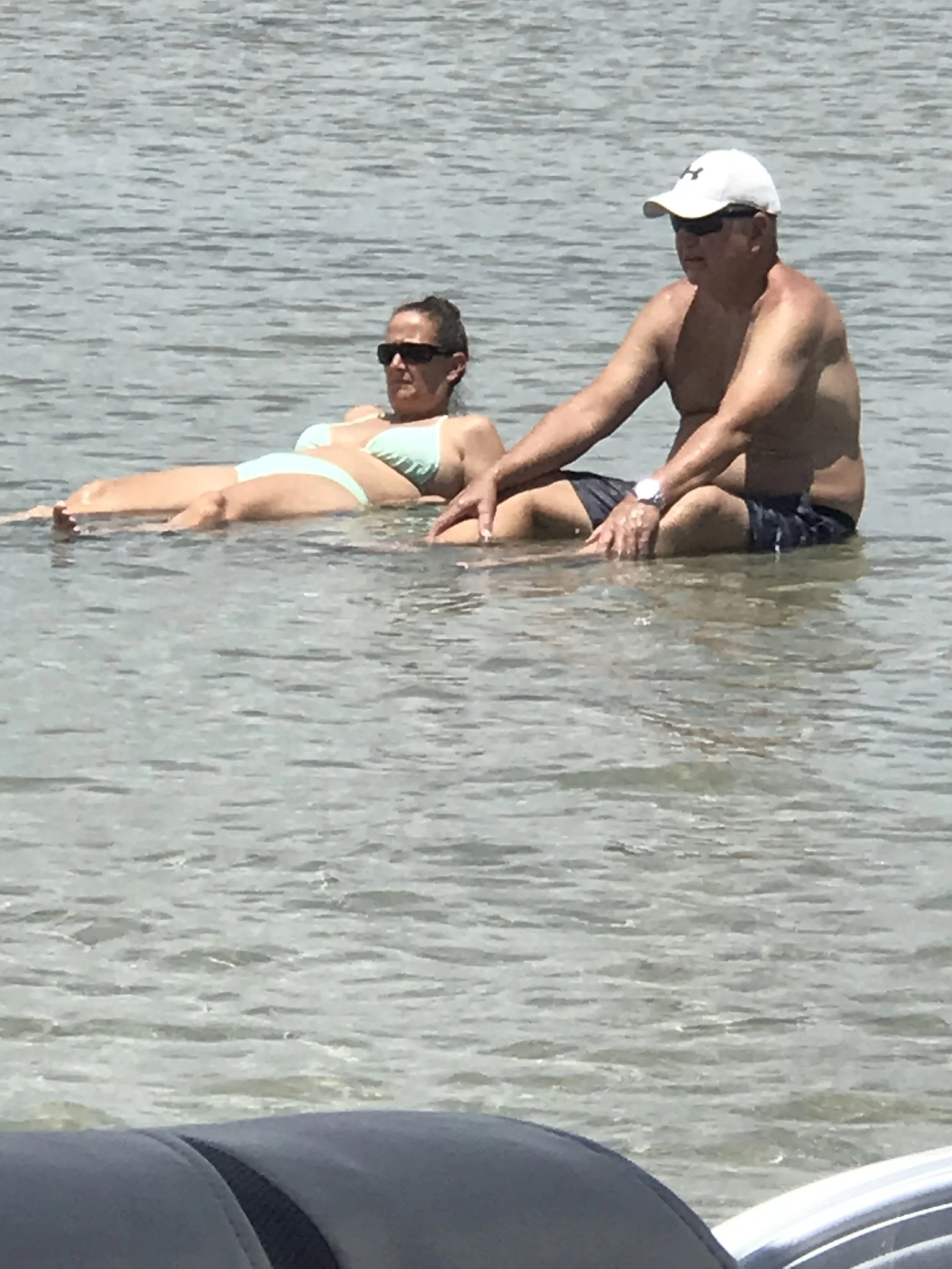 A man and woman sitting in shallow water at the beach, both wearing sunglasses and the man wearing a cap, with a car visible in the foreground.