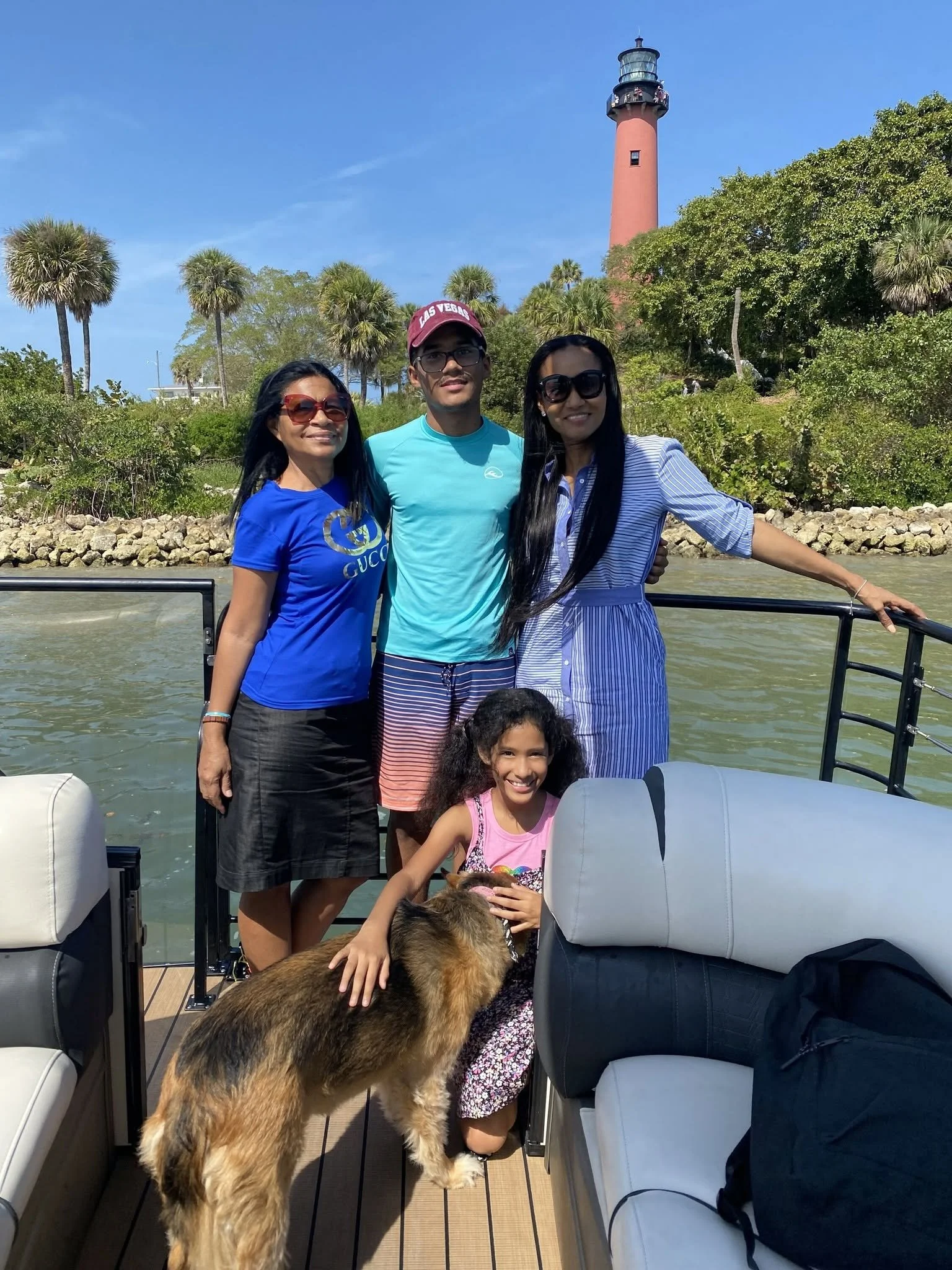 Group of people on a boat near a lighthouse with trees and blue sky in the background. There are three women and one young girl, with a dog visible in the foreground.