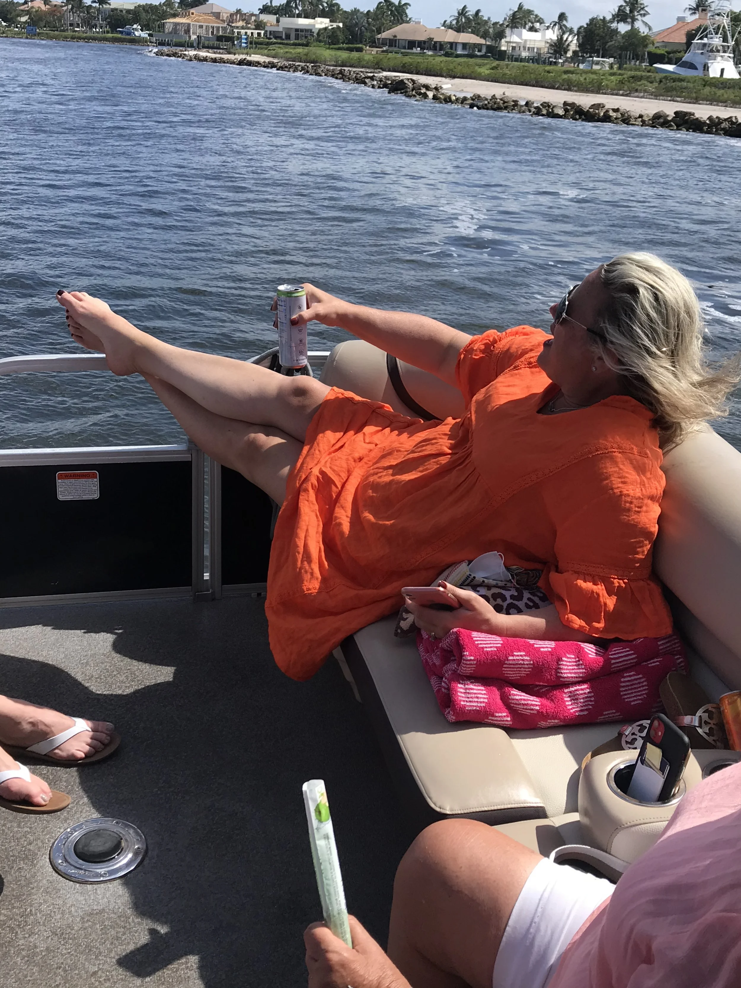 A woman relaxing on a boat near the water, holding a can of drink, with other people's legs and feet visible nearby. The background shows houses and palm trees along the shoreline.