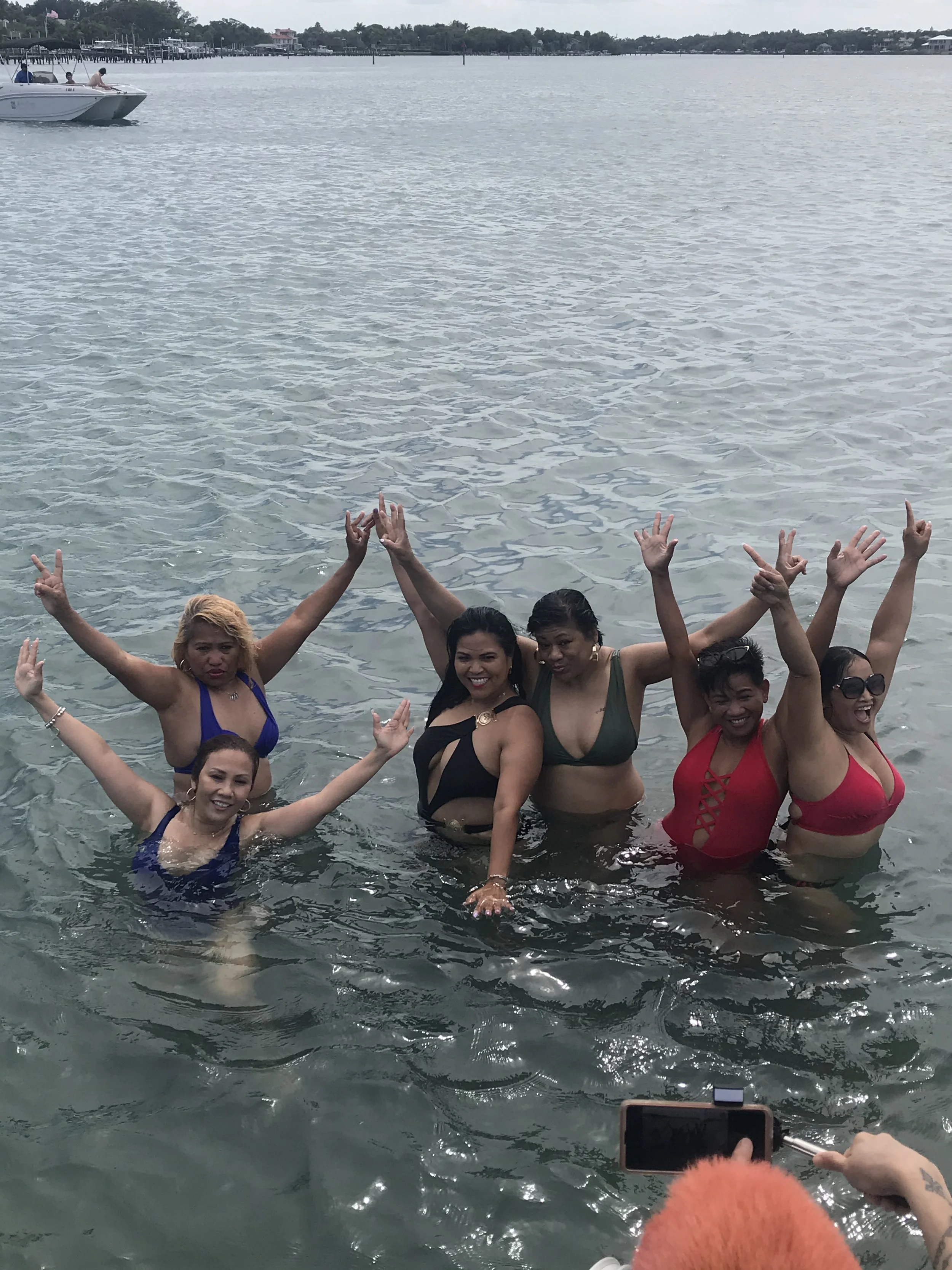 Group of women in swimsuits standing in water near a dock, raising their hands and smiling at the camera with boats and a shoreline in the background.