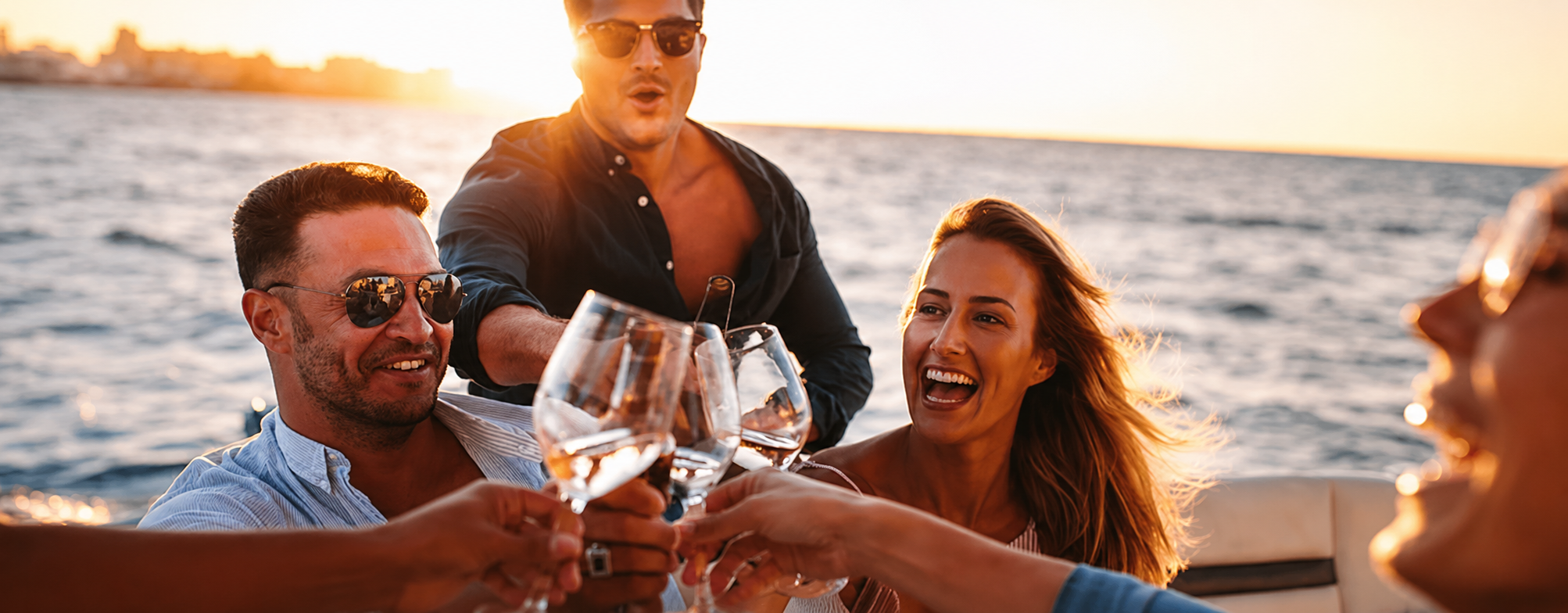 Group of friends enjoying drinks on a boat at sunset, smiling and toasting with wine glasses, with water and city skyline in the background.