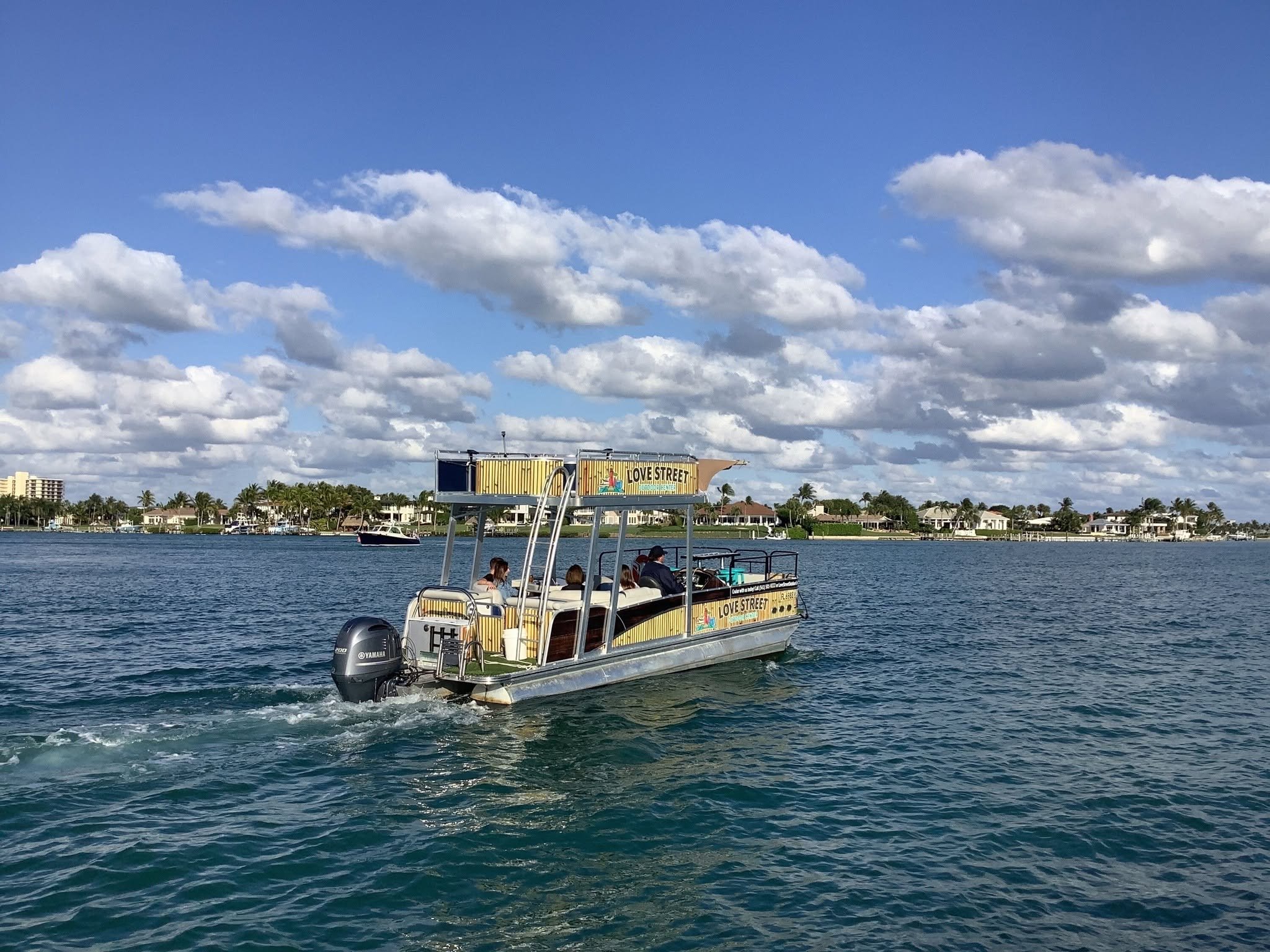 A boat with passengers on a body of water, with a billboard sign reading 'Love Street' in the background and residential buildings along the shoreline, under a partly cloudy sky.