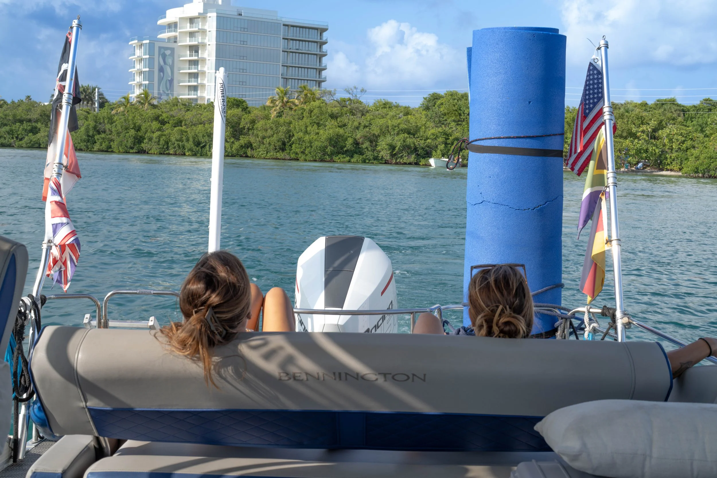 View from the back of a boat with three children sitting and facing the water, with lounge cushions. There is a blue foam post, flags, and an outboard motor visible. The water is calm with a wooded shoreline and a modern building in the background.