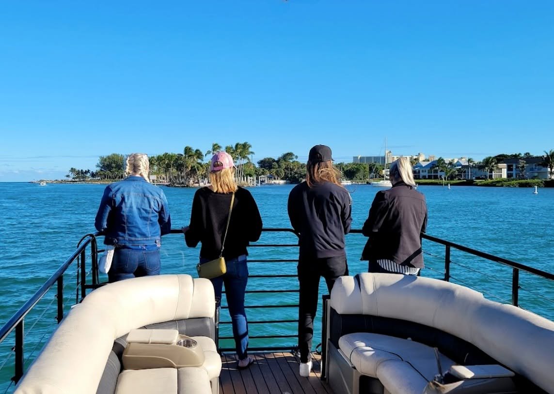 Four women standing on the deck of a boat, looking out at a waterway with palm trees and buildings in the background.