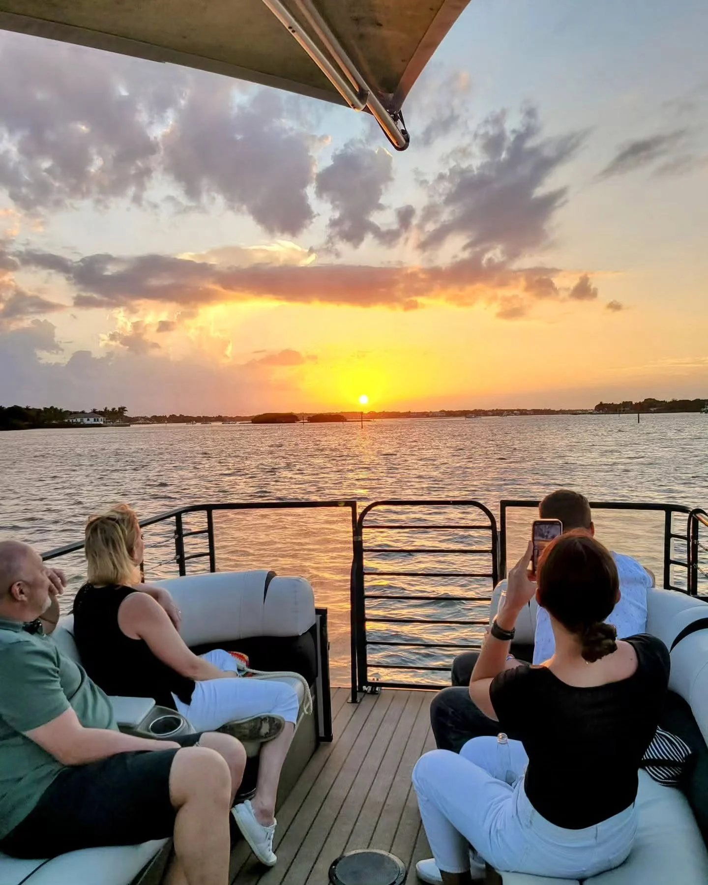 People sitting on a boat deck during sunset, with water and sky in the background and some taking photos.