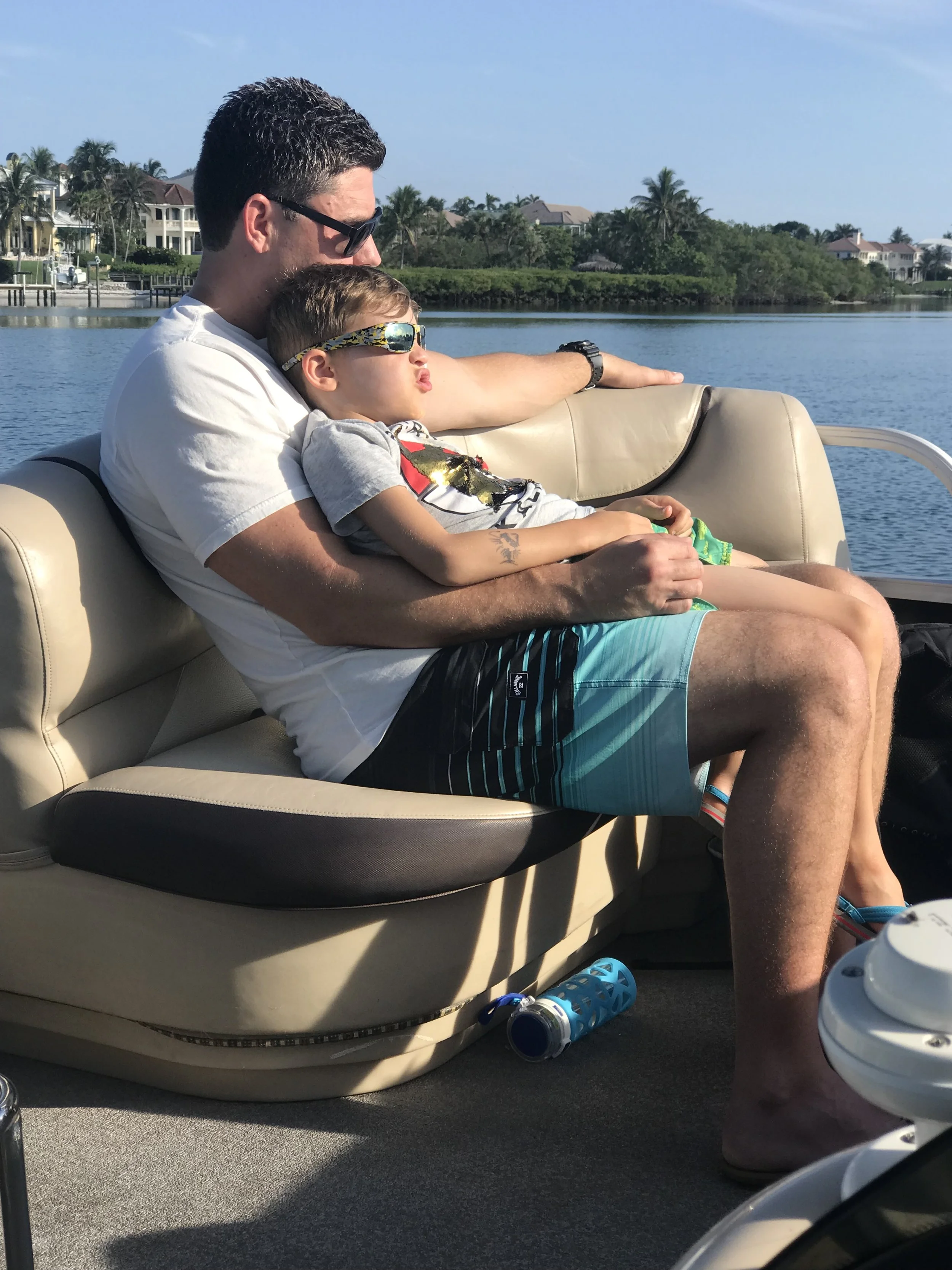 A man and a young boy sitting on a boat seat near a body of water with houses and palm trees in the background, wearing sunglasses, enjoying a sunny day.