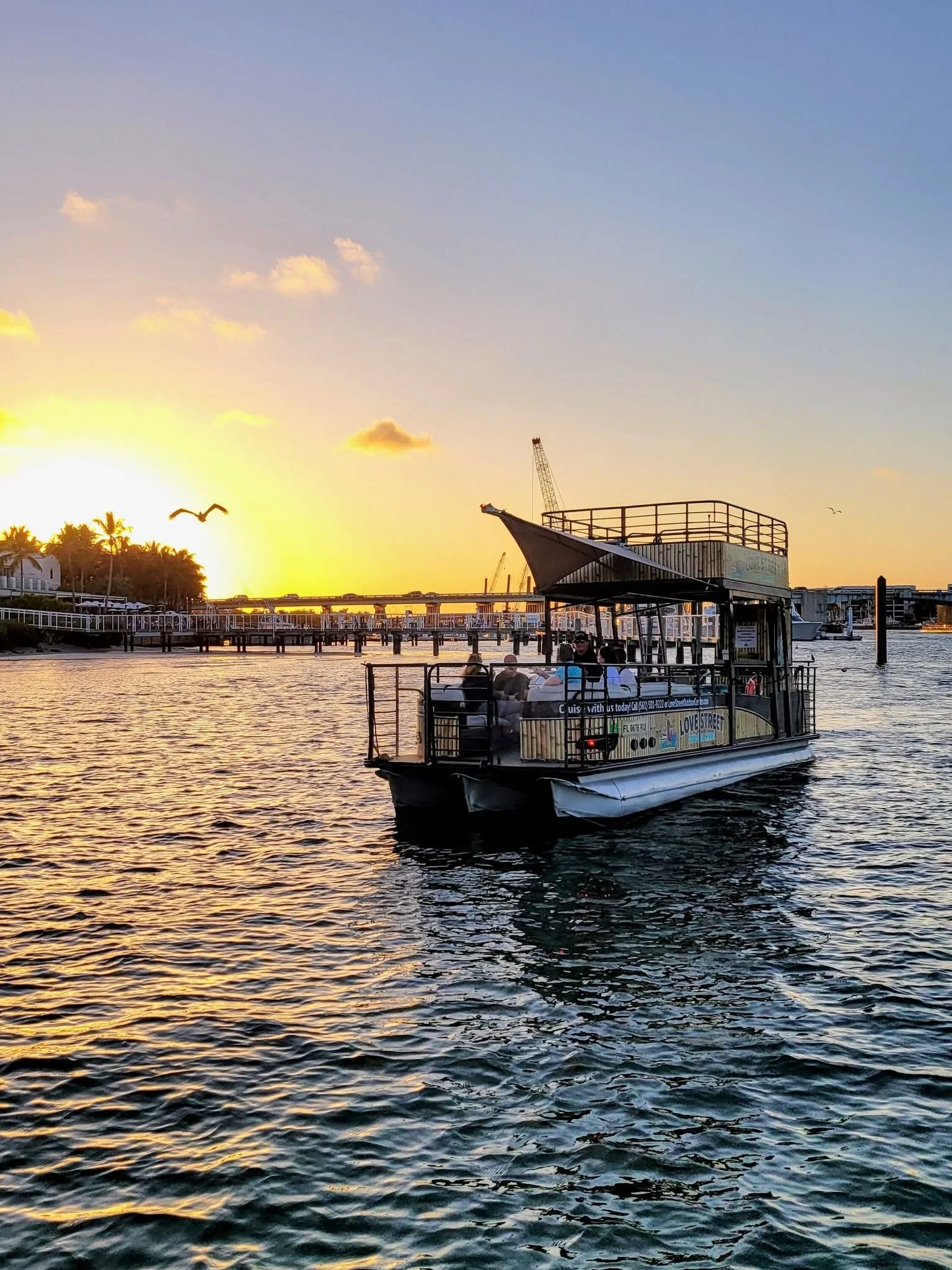 A boat on the water during sunset with a bridge and palm trees in the background and a bird flying overhead.