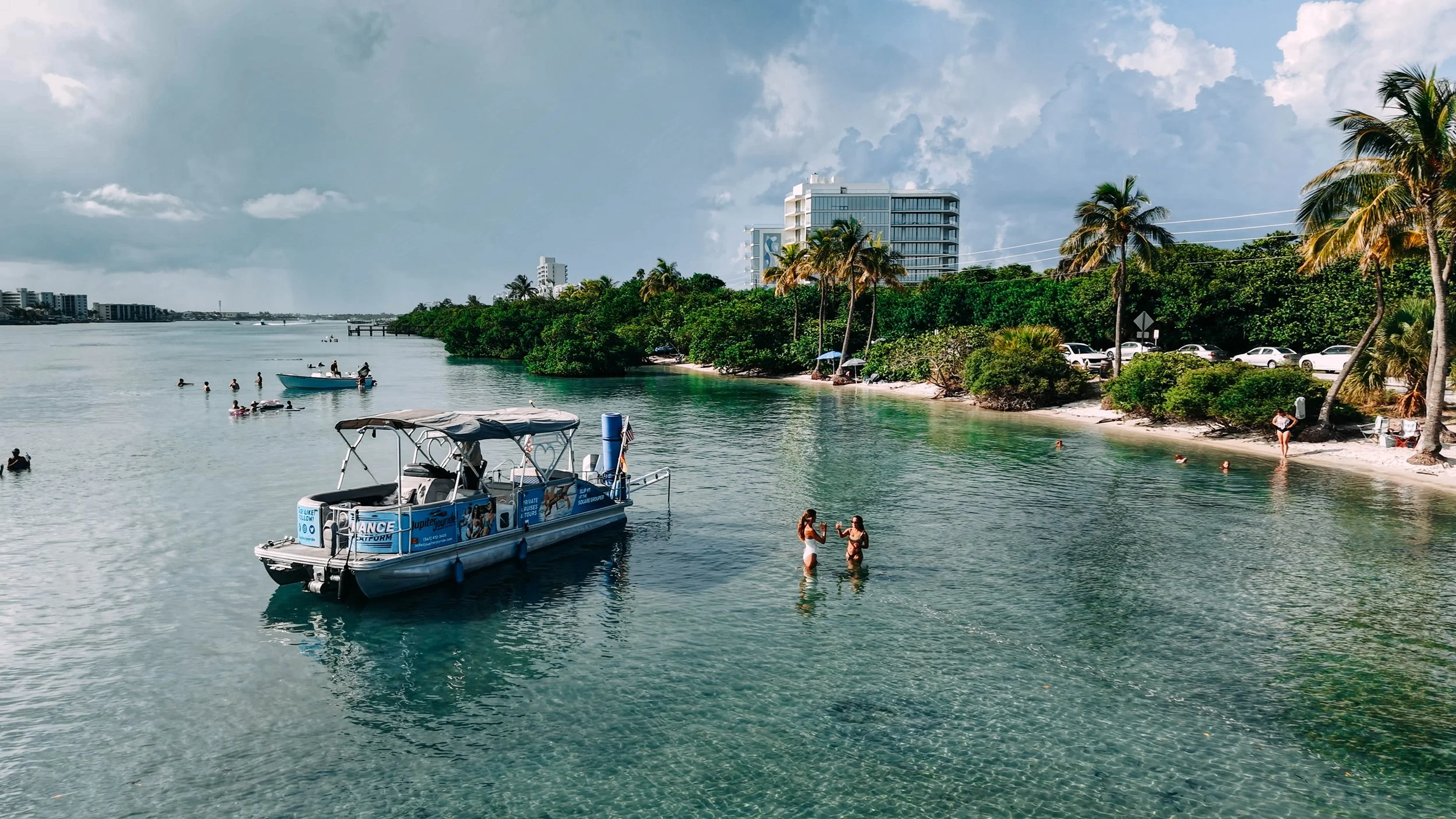 People swimming and relaxing along a tropical shoreline with palm trees, a boat, and modern high-rise buildings in the background.