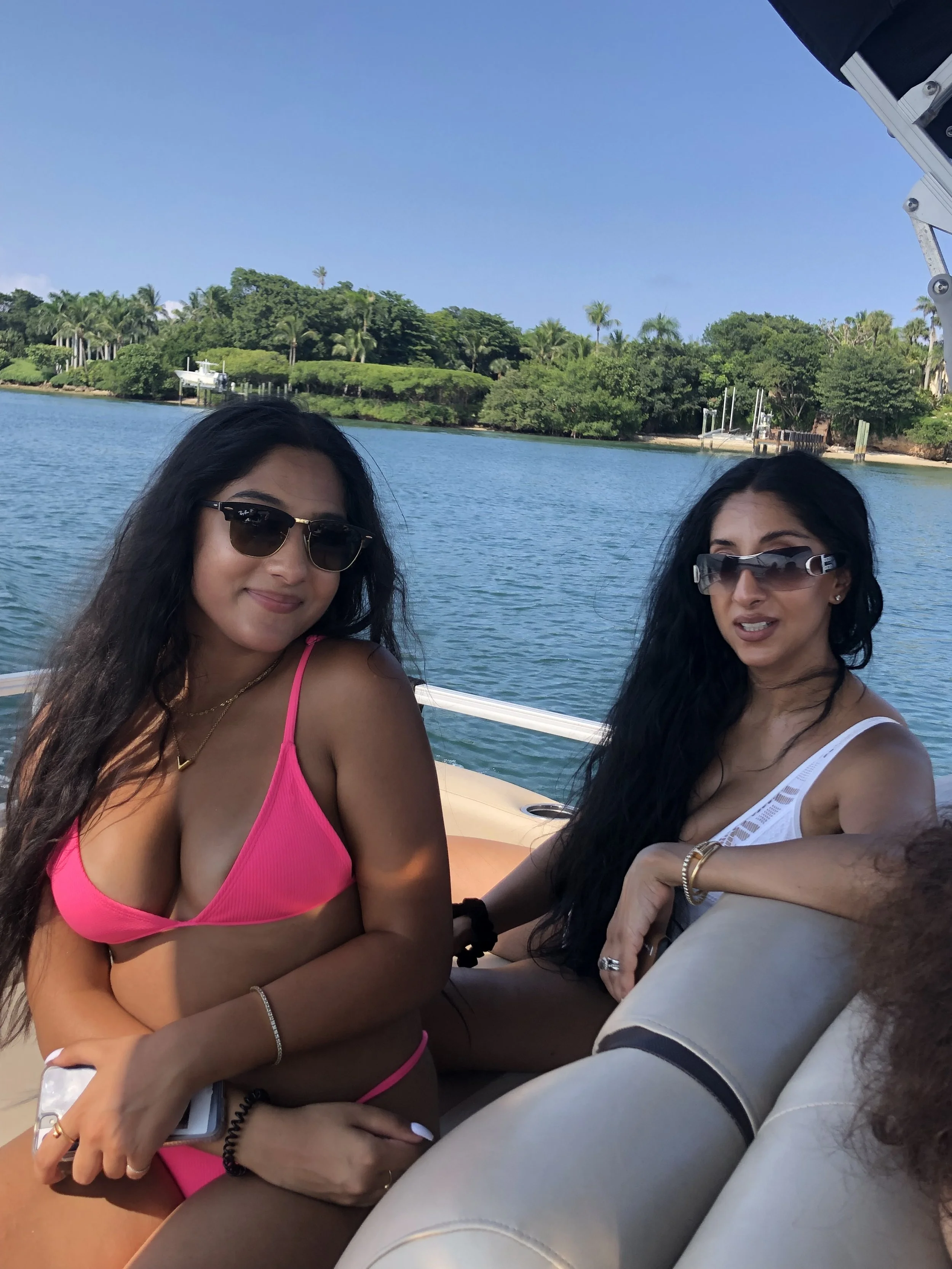 Two women sitting on a boat with a waterway and green trees in the background, wearing sunglasses and casual summer clothes.