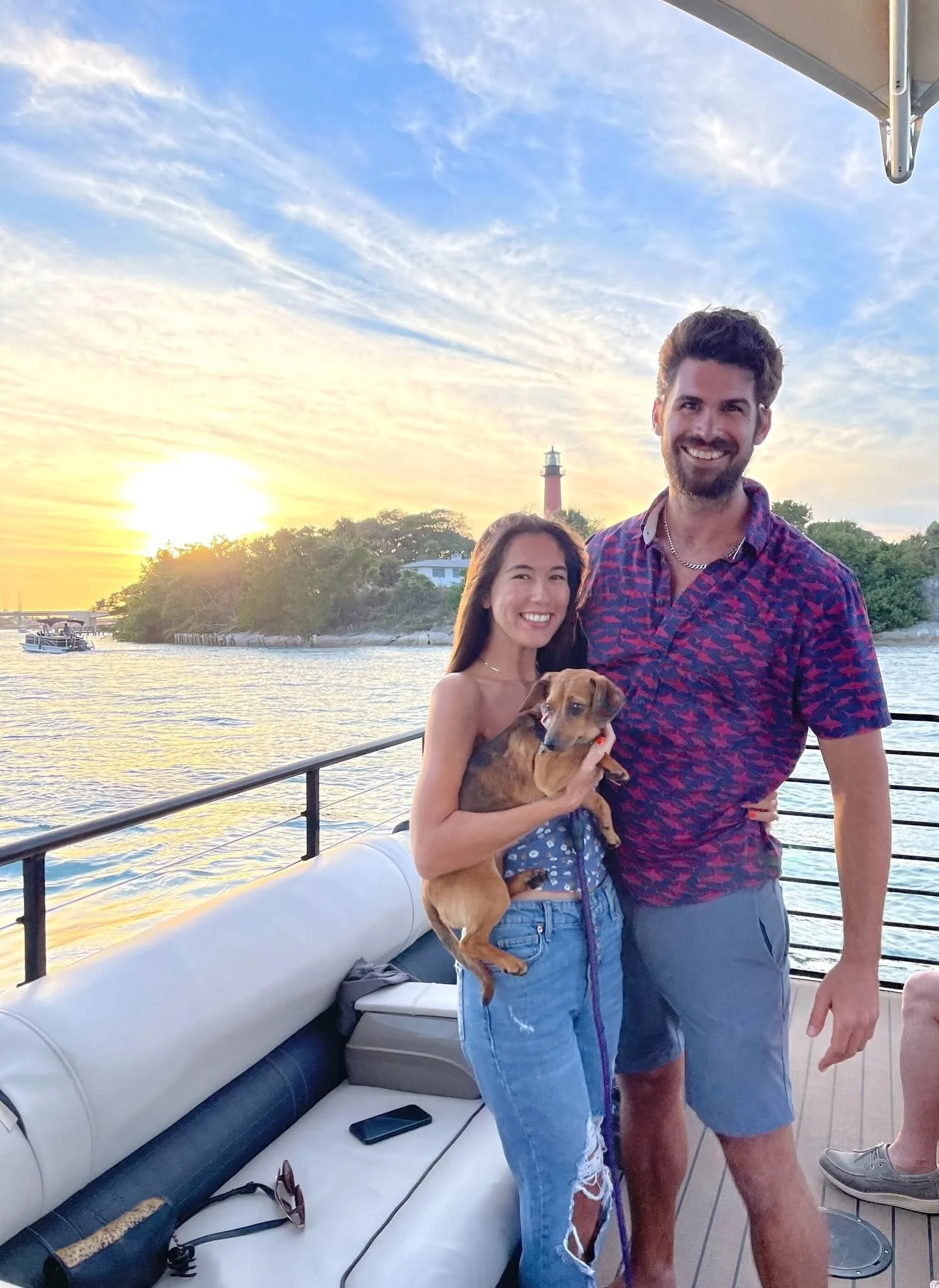A young woman and man smiling on a boat at sunset, holding a small dog, with water and a lighthouse in the background.