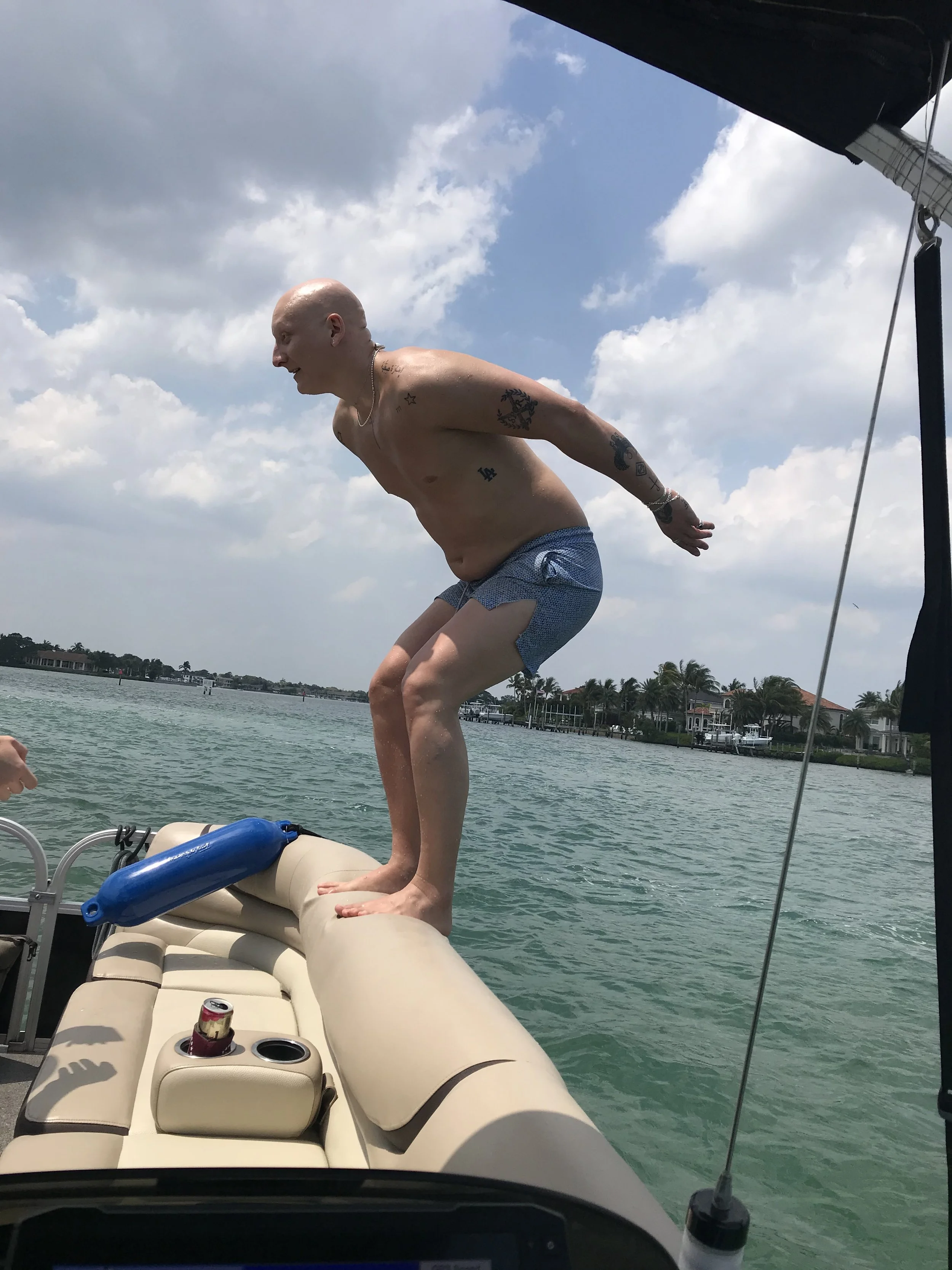 A bald man with tattoos on his arms and chest, wearing swimming trunks, is balancing on the cushion of a boat seat over water. The boat has a beige interior with a cup holder containing a soda can. The sky is partly cloudy and houses are visible on t