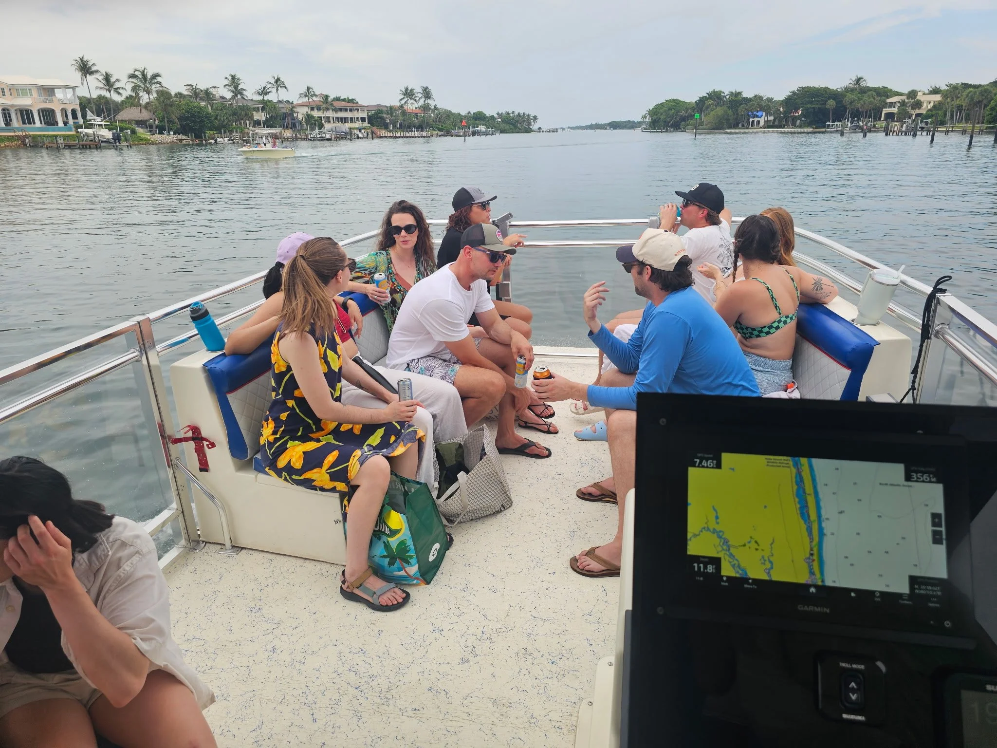 A group of people sitting on a boat's deck overlooking a waterway with houses and palm trees along the banks, some drinking beverages and chatting, with a GPS device showing navigation data in the foreground.