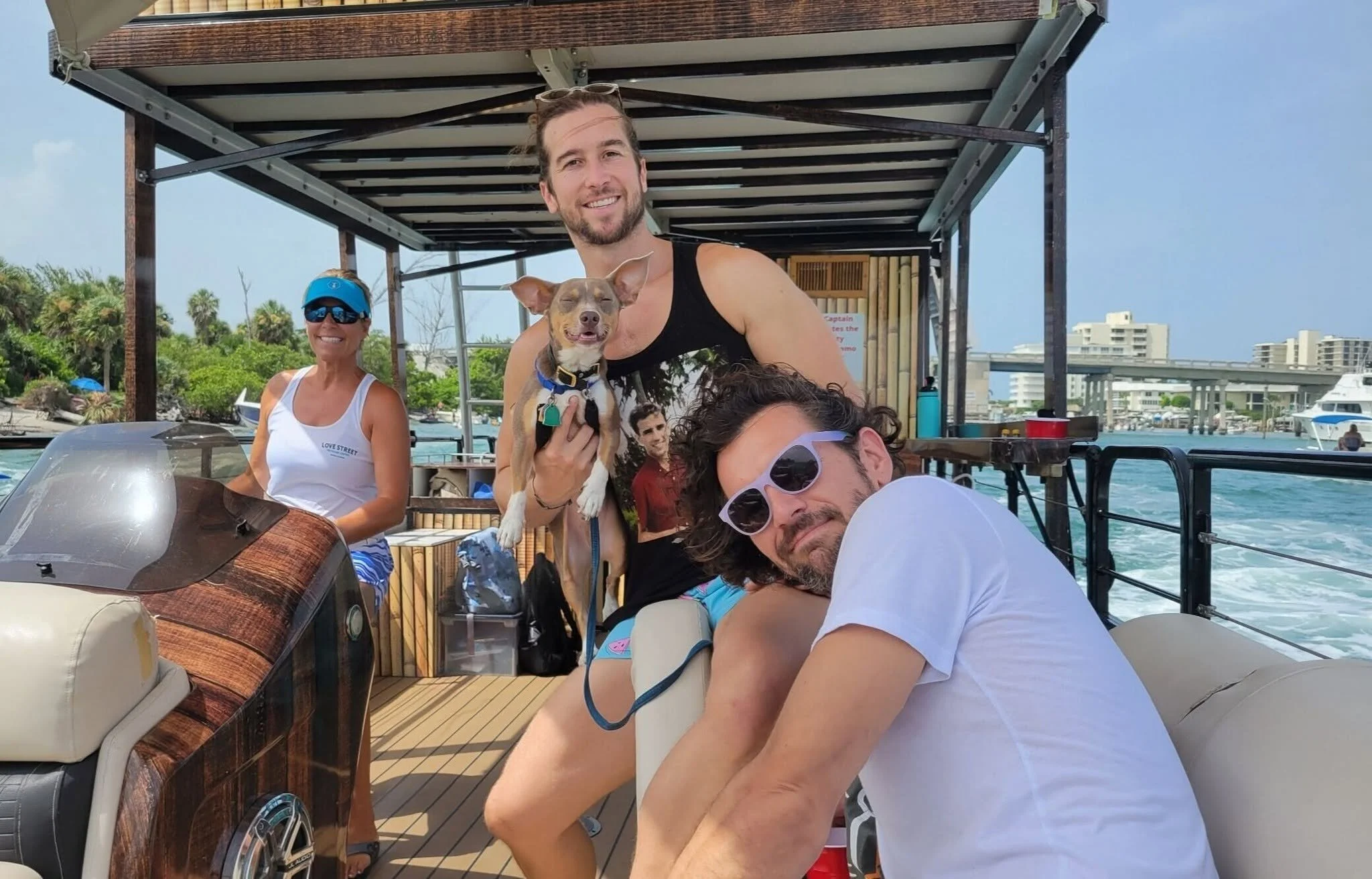 Group of people enjoying a boat ride on the water, with a man, woman, and dog in the foreground, and city buildings and boats in the background.