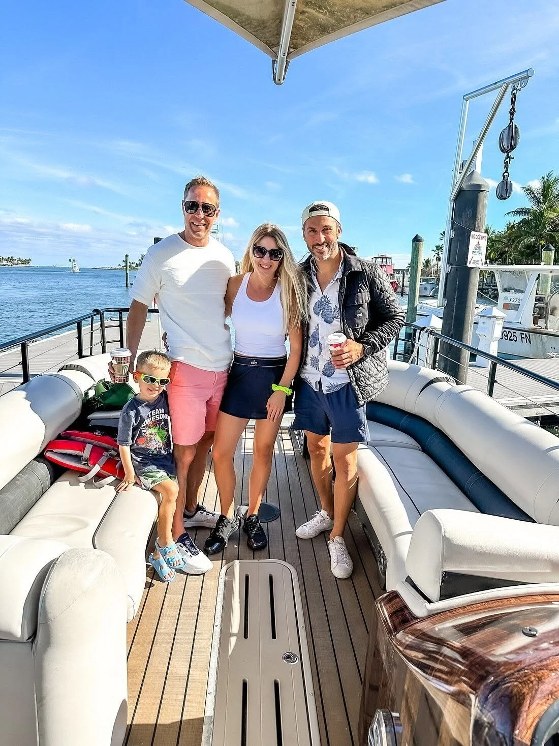 A group of four adults and one child on a boat docked at a marina, smiling for the camera with water and boats in the background on a sunny day.
