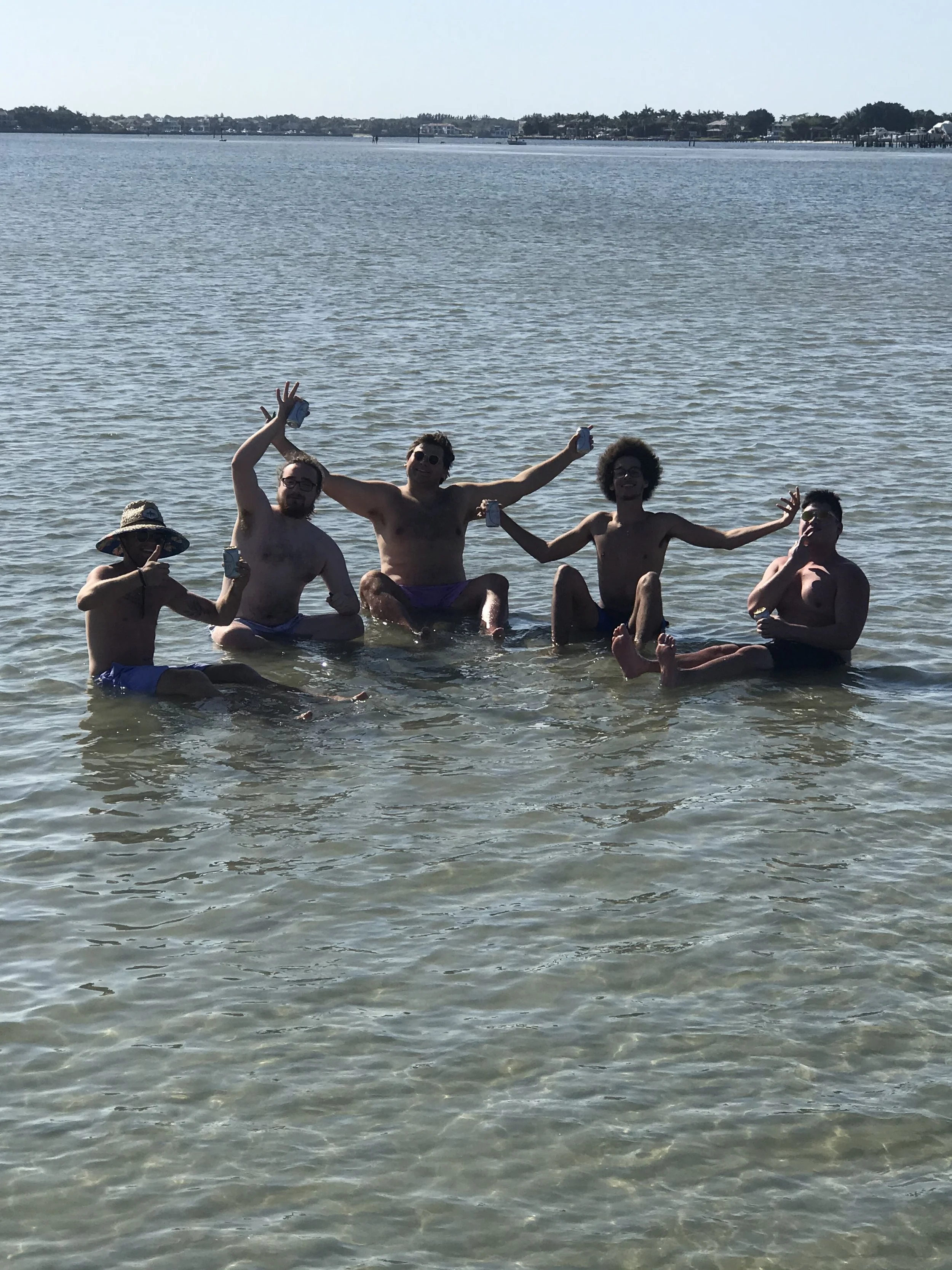 Five shirtless men sitting and standing in shallow water by a shoreline, holding cans, with a calm body of water and distant buildings and trees in the background.