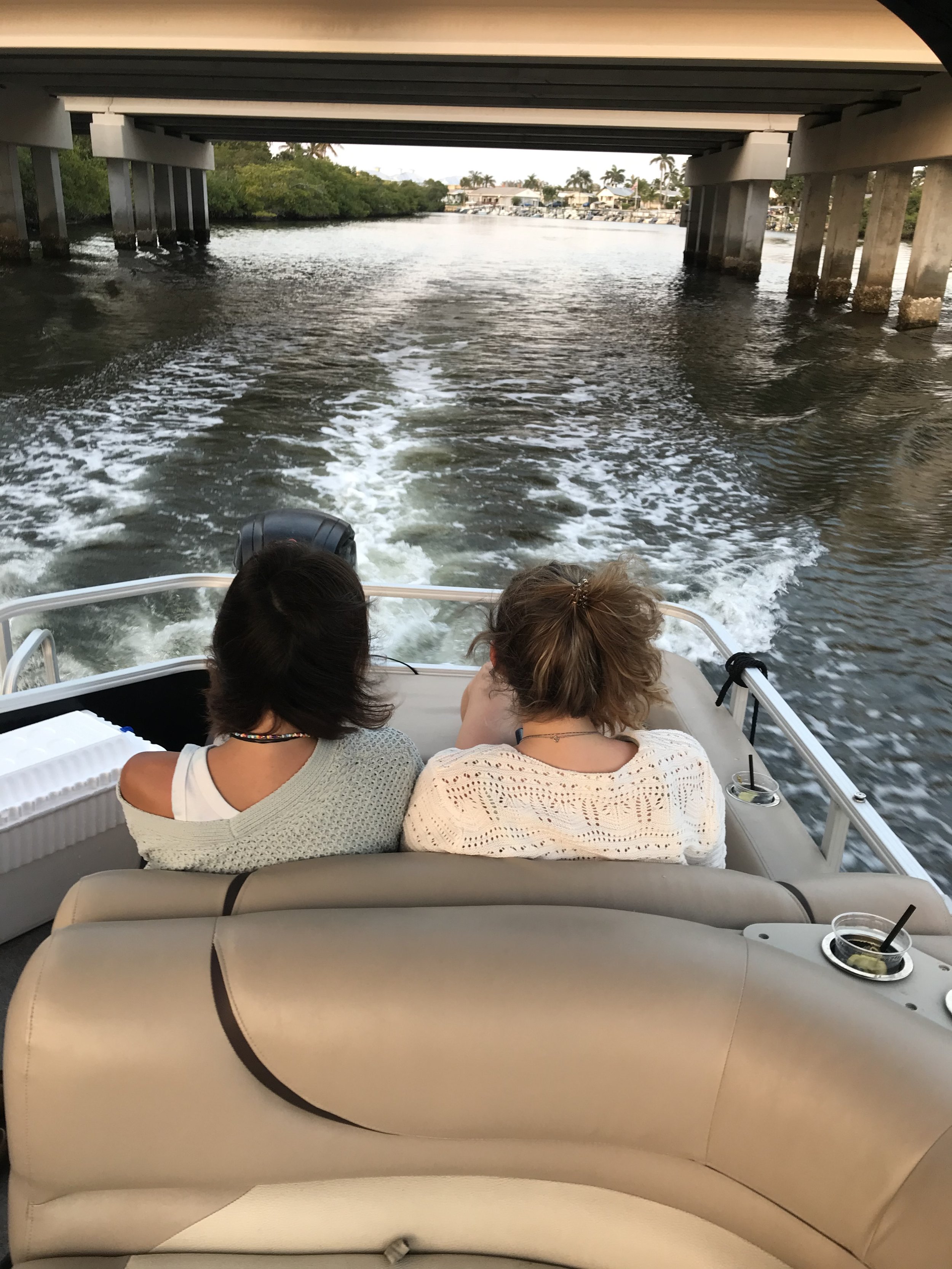 People sitting on a boat, view of water and a bridge overhead, with houses and boats in the background.