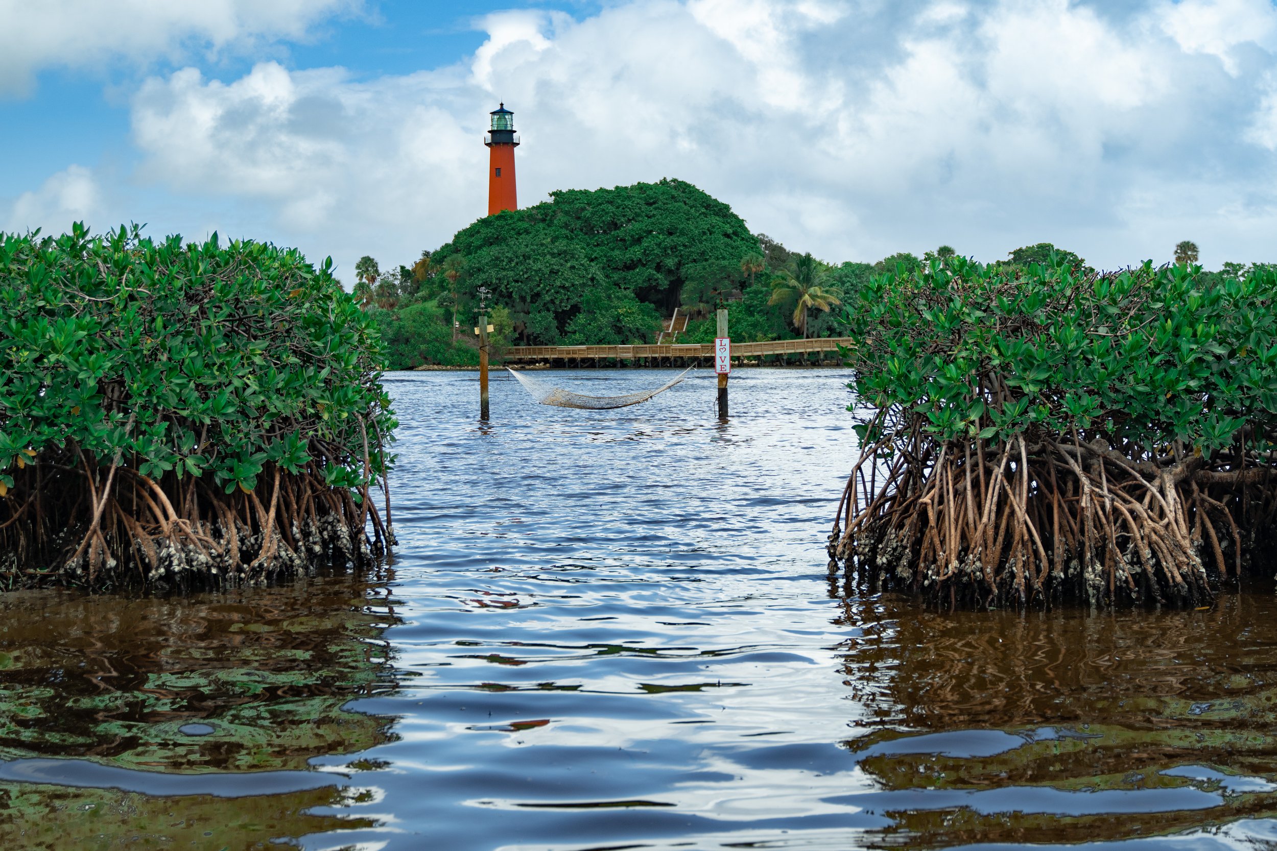 Tidal mangroves in the foreground with water, a small pier, a lush green island with a lighthouse on top, and a partly cloudy sky in the background.