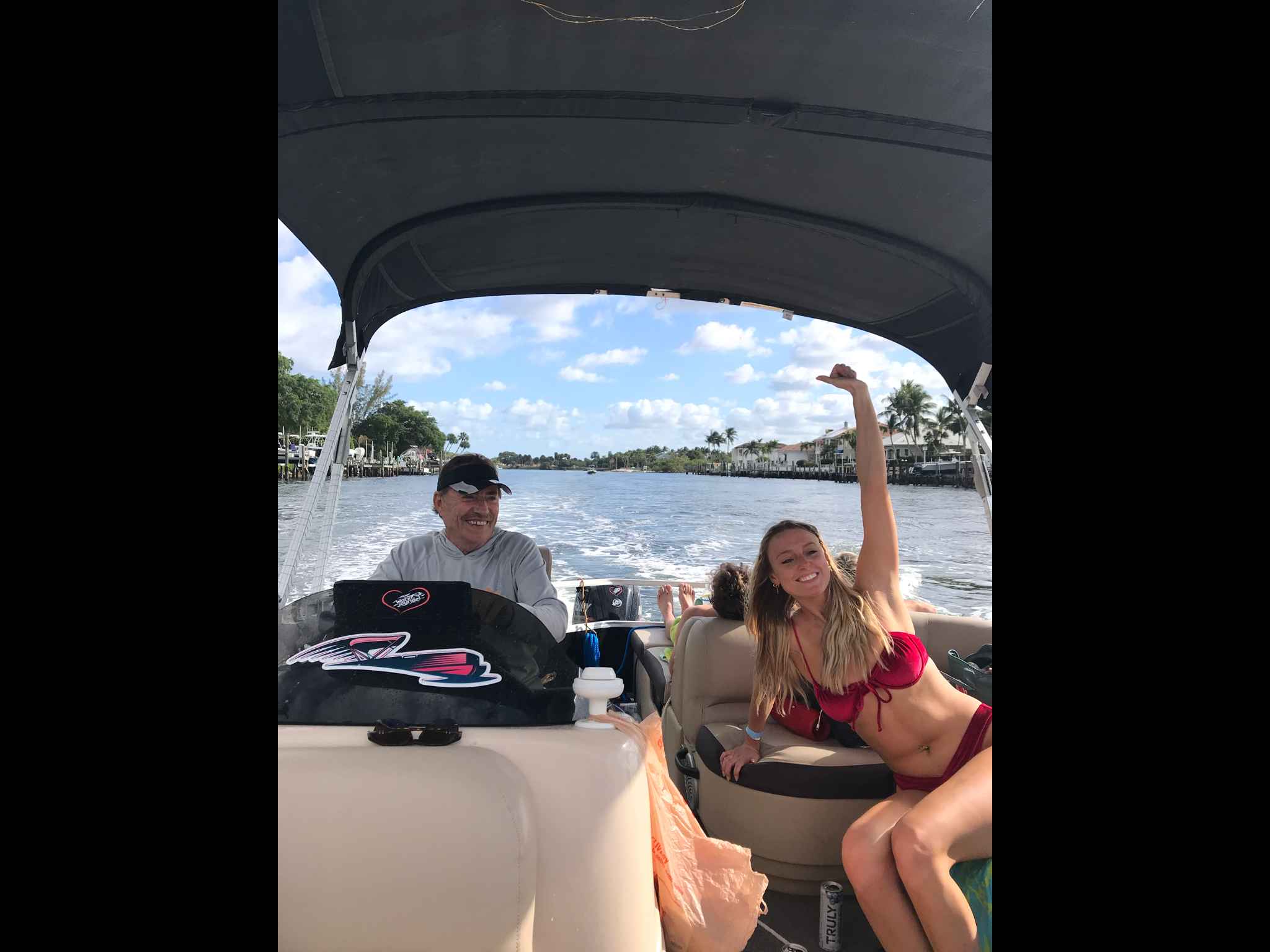 Two people enjoying a boat ride on a canal with a waterfront neighborhood in the background. One woman in a red bikini is smiling and waving, and an older man is sitting next to her, smiling. The boat has a canopy, and the waterway is lined with palm