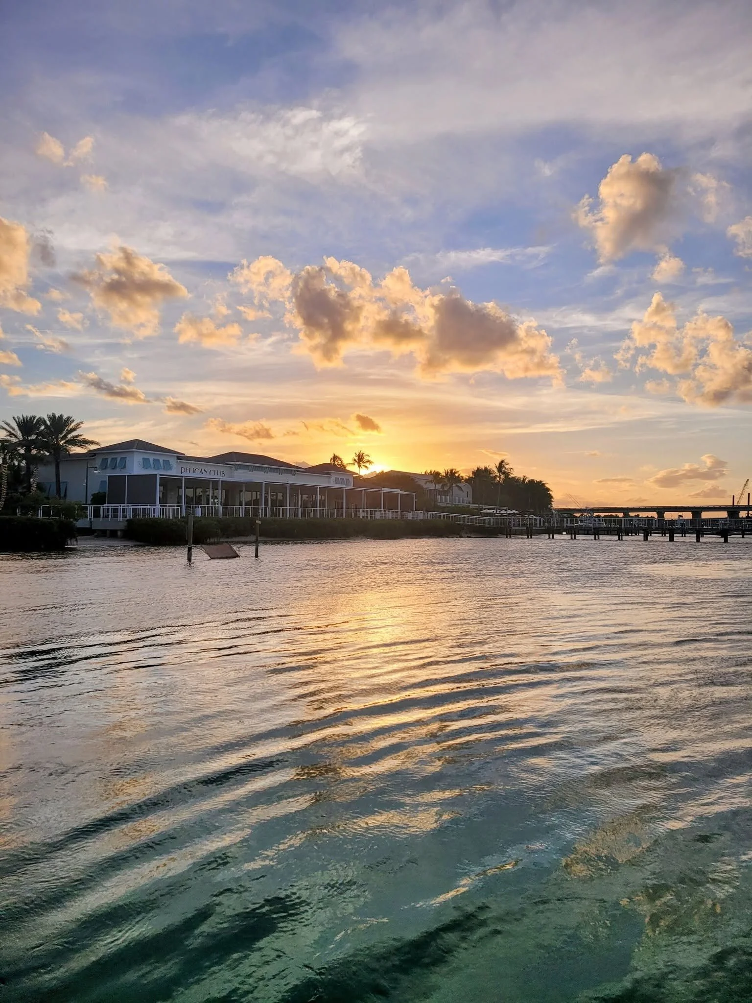 Sunset over a body of water with clouds in the sky and buildings and palm trees along the shoreline.