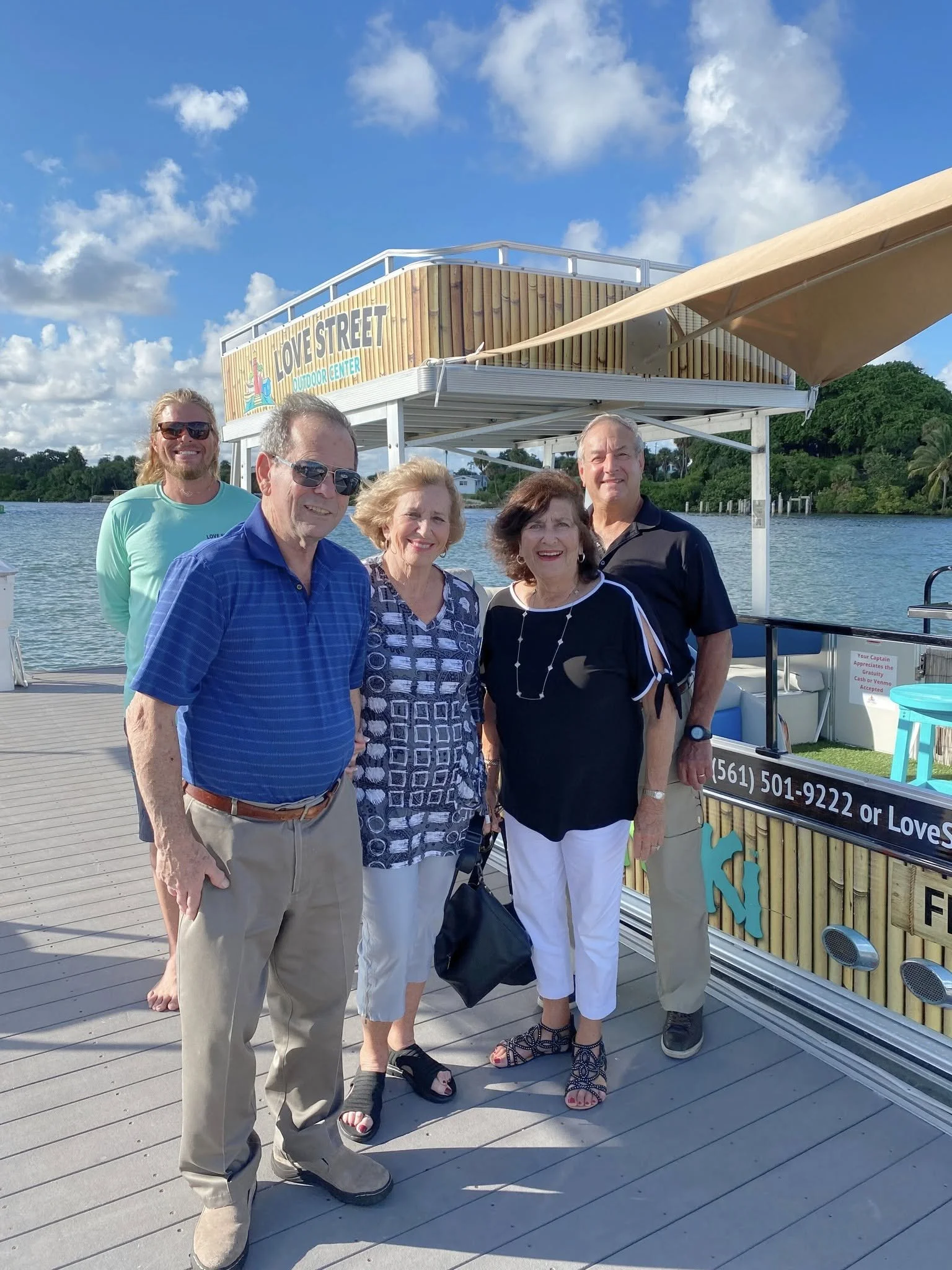 Group of five people standing on a dock near water with a boat and a wooden structure that reads 'Loves Street Outdoor Center' in the background.