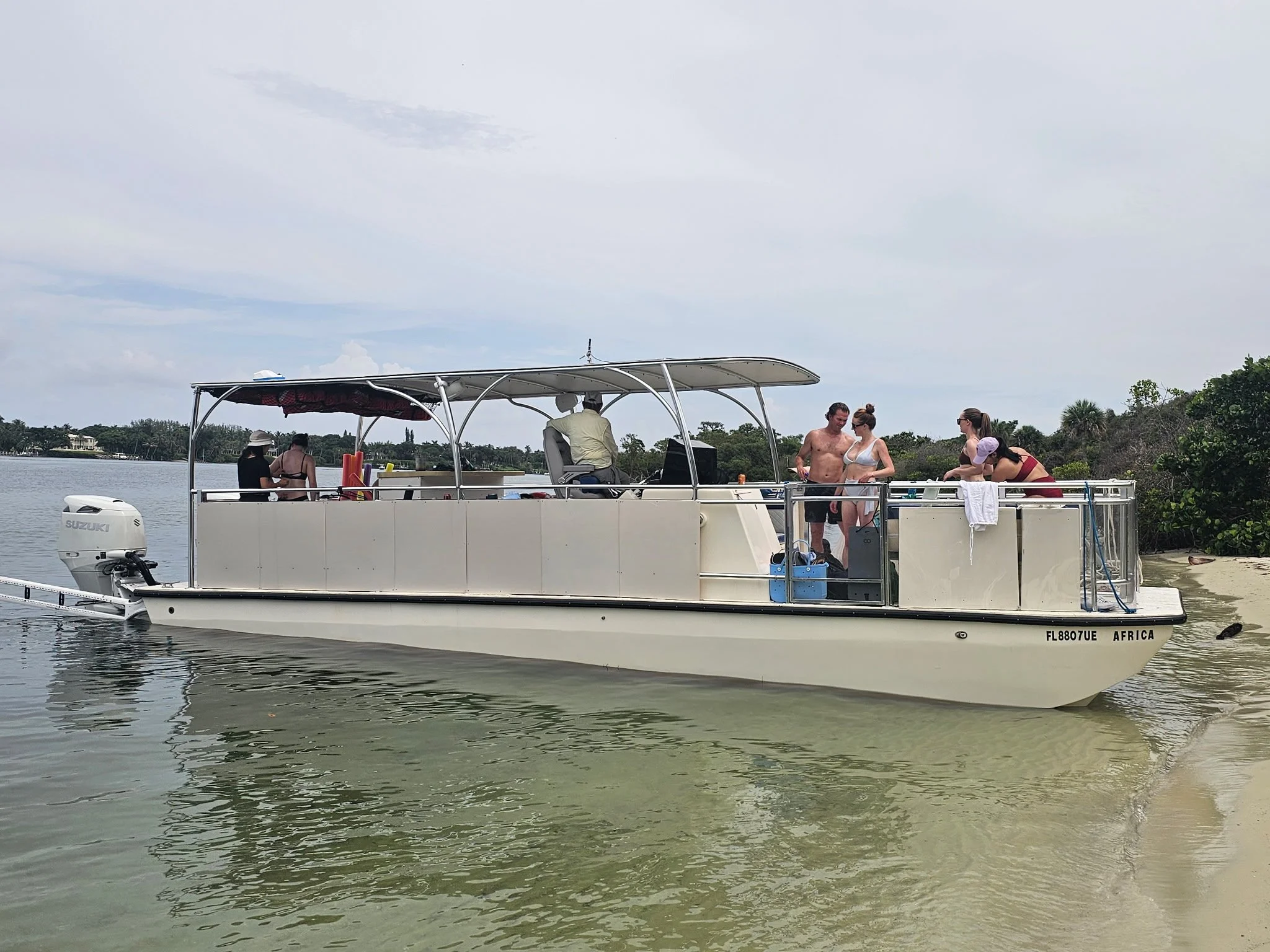A white boat with a Suzuki outboard motor is on water with four people onboard. Two women and a man are near the front, with one woman wearing a purple hat and others in swimsuits. A man is steering at the helm, and two women are at the back. The boat has a shade canopy and is near some greenery and water. Clouds are visible in the sky.