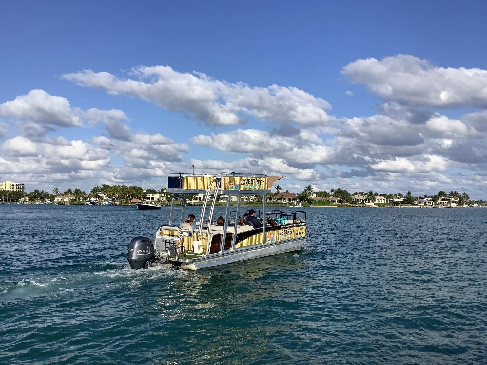 A boat named Love Street with people on board, cruising on a waterway with houses and palm trees in the background, under a partly cloudy sky.