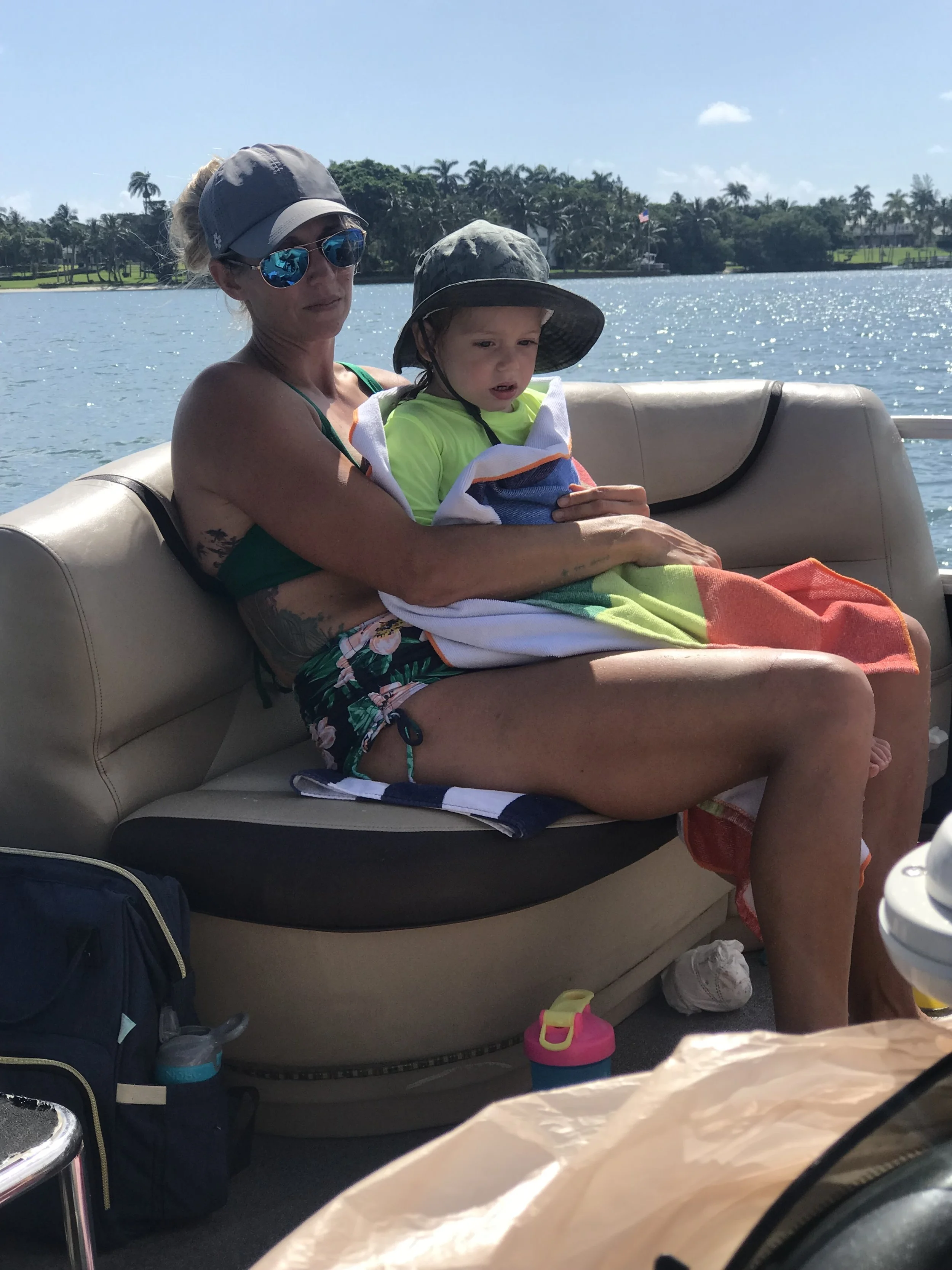 A woman and a young boy sitting on a boat during daytime, with trees and water in the background.