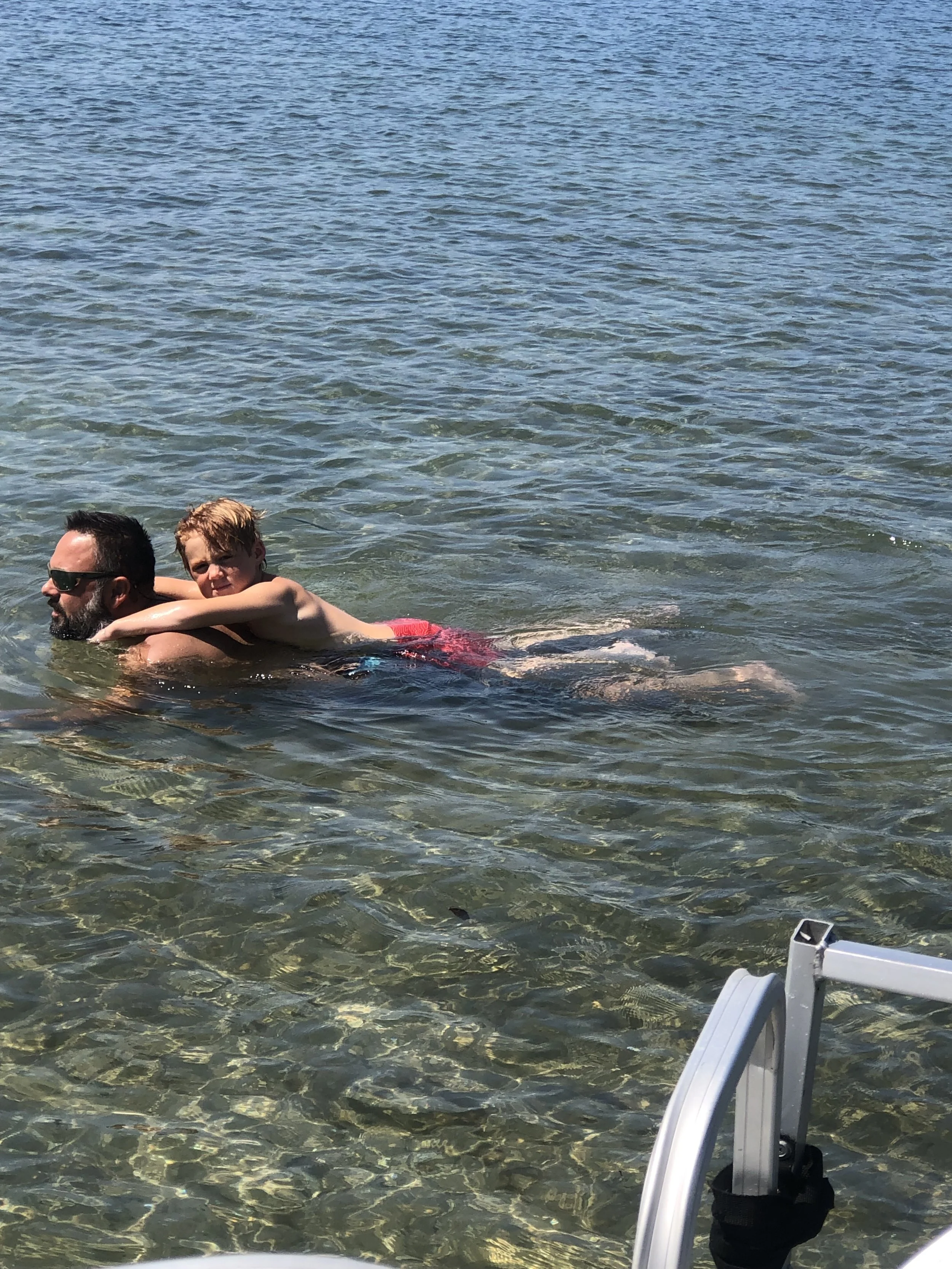 A man and a young boy swim together in clear, shallow water near the shore. The man wears sunglasses, and the boy has red hair. The water is calm and transparent, revealing the sandy bottom.