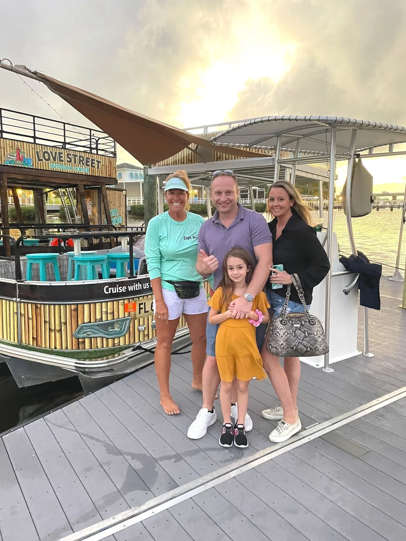 A group of five people, including three women, an older woman with a visored cap, a man, and a young girl, are smiling and posing on a boat dock with a boat and water in the background at sunset.