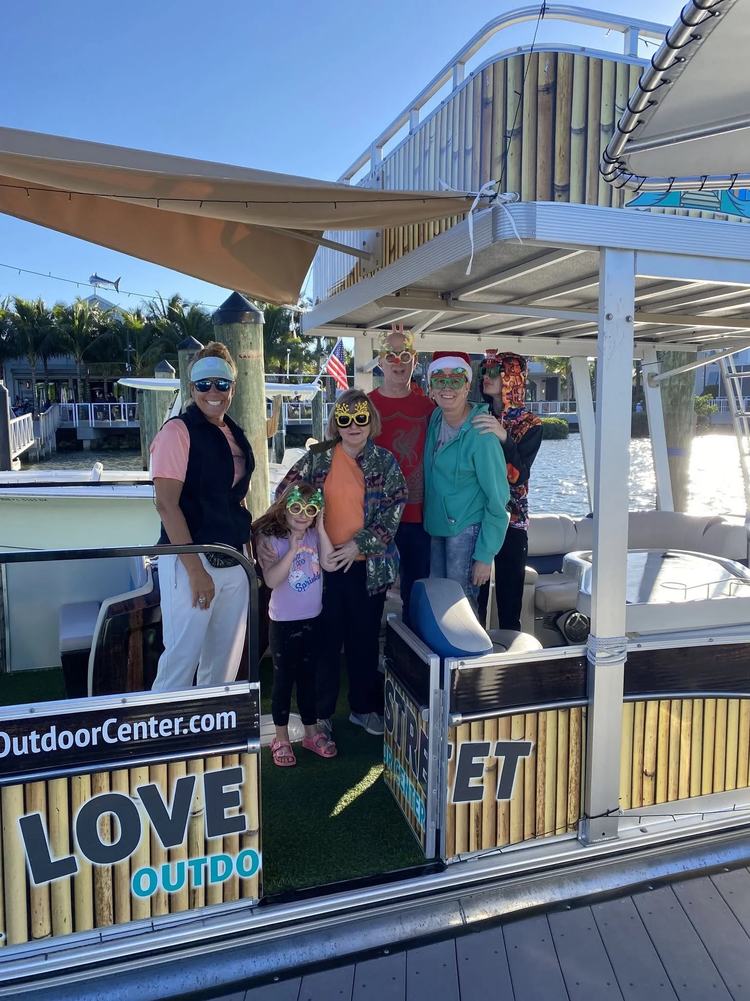 Group of six people, including four women and two children, wearing holiday-themed sunglasses and costumes, standing on a dock by a boat with a sign that reads 'LOVE OUTDOOR' and 'LOVE'. The group is smiling and posing for a photo with water in the b