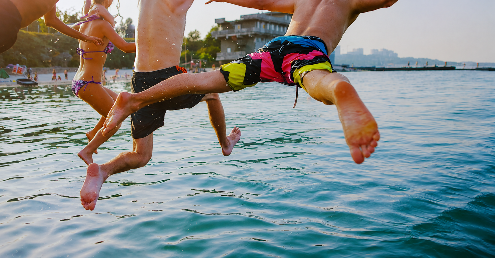 People jumping into the water at a beach during sunset, wearing swimwear, with a cityscape and trees in the background.
