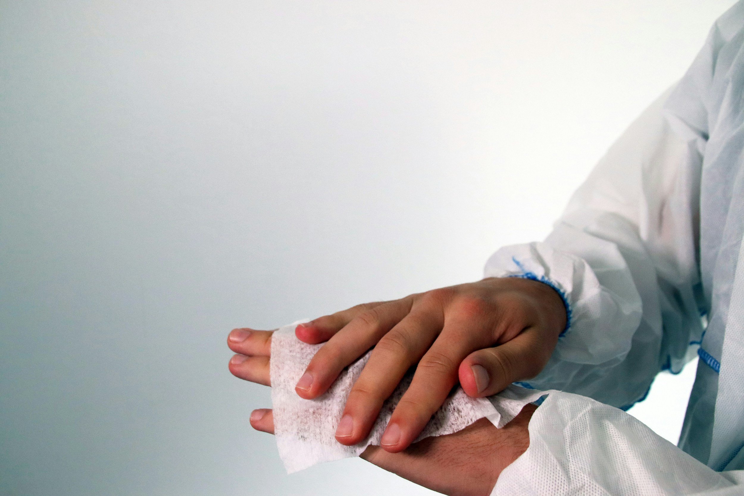 A healthcare professional in a white protective gown holds a patient's hand, which is wrapped with a medical gauze or bandage, against a plain light-colored background.