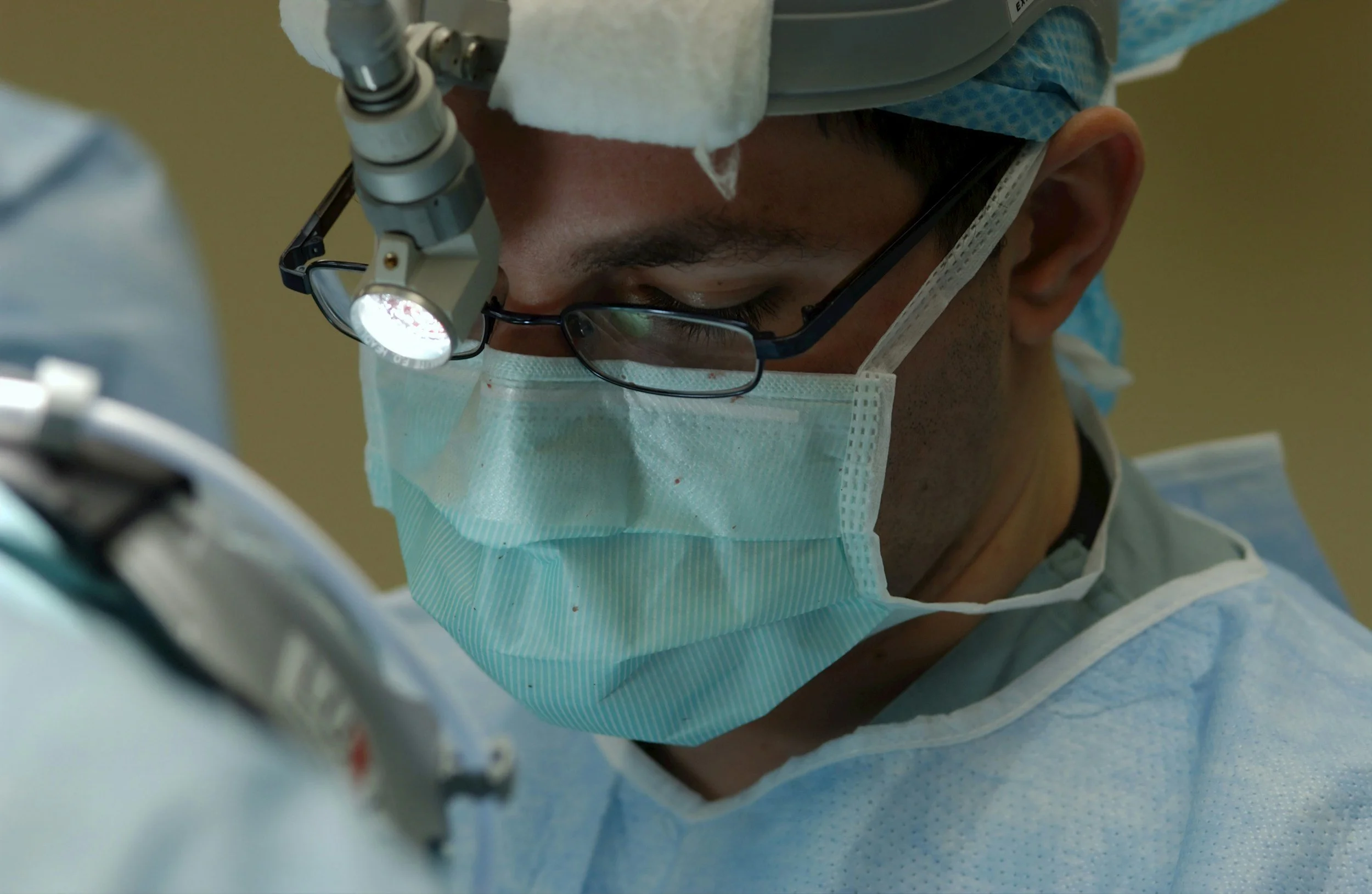 A surgeon wearing glasses and a surgical mask, working under a bright light in an operating room.
