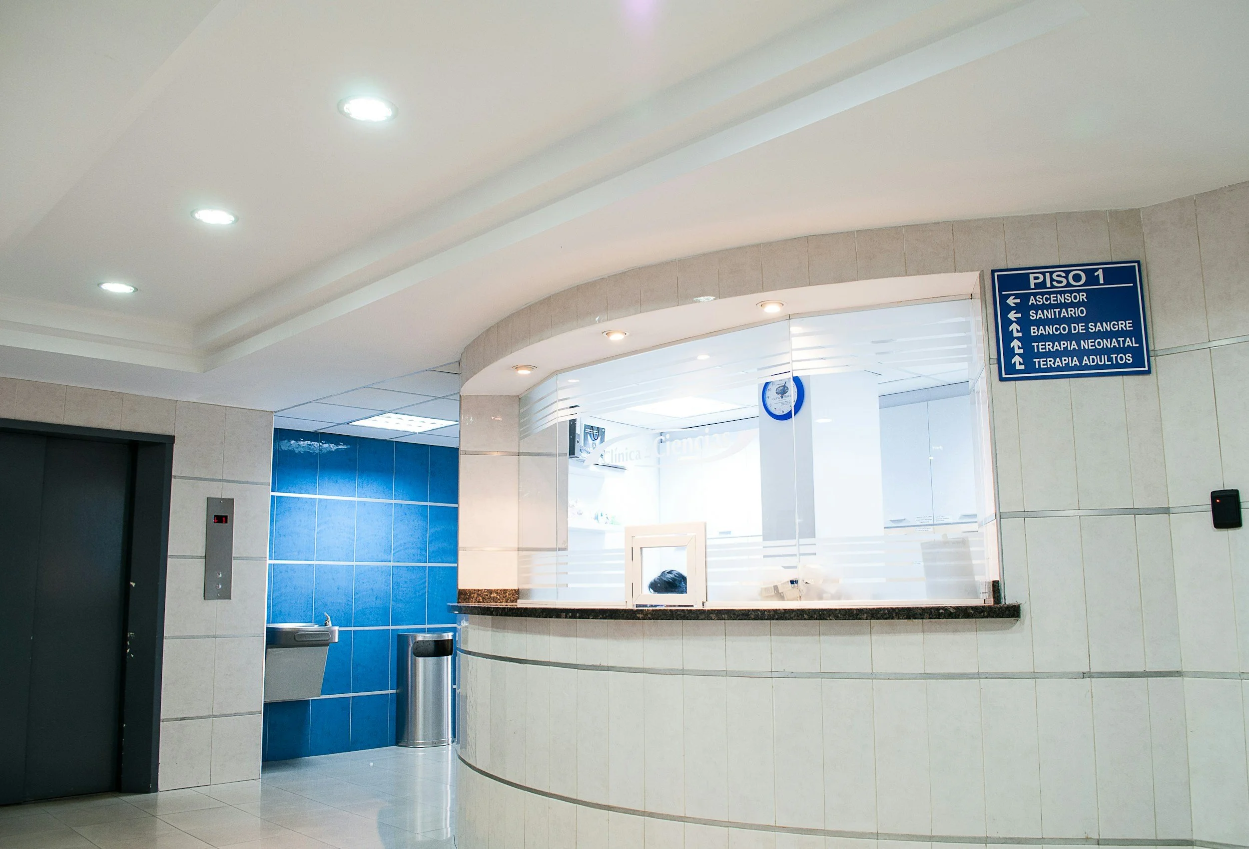 Interior view of a hospital or clinic reception area with a blue sign indicating the first floor and directions to elevator, restroom, blood bank, neonatal therapy, and adult therapy. The area features tiled walls, ceiling lights, a counter, and an elevator door.