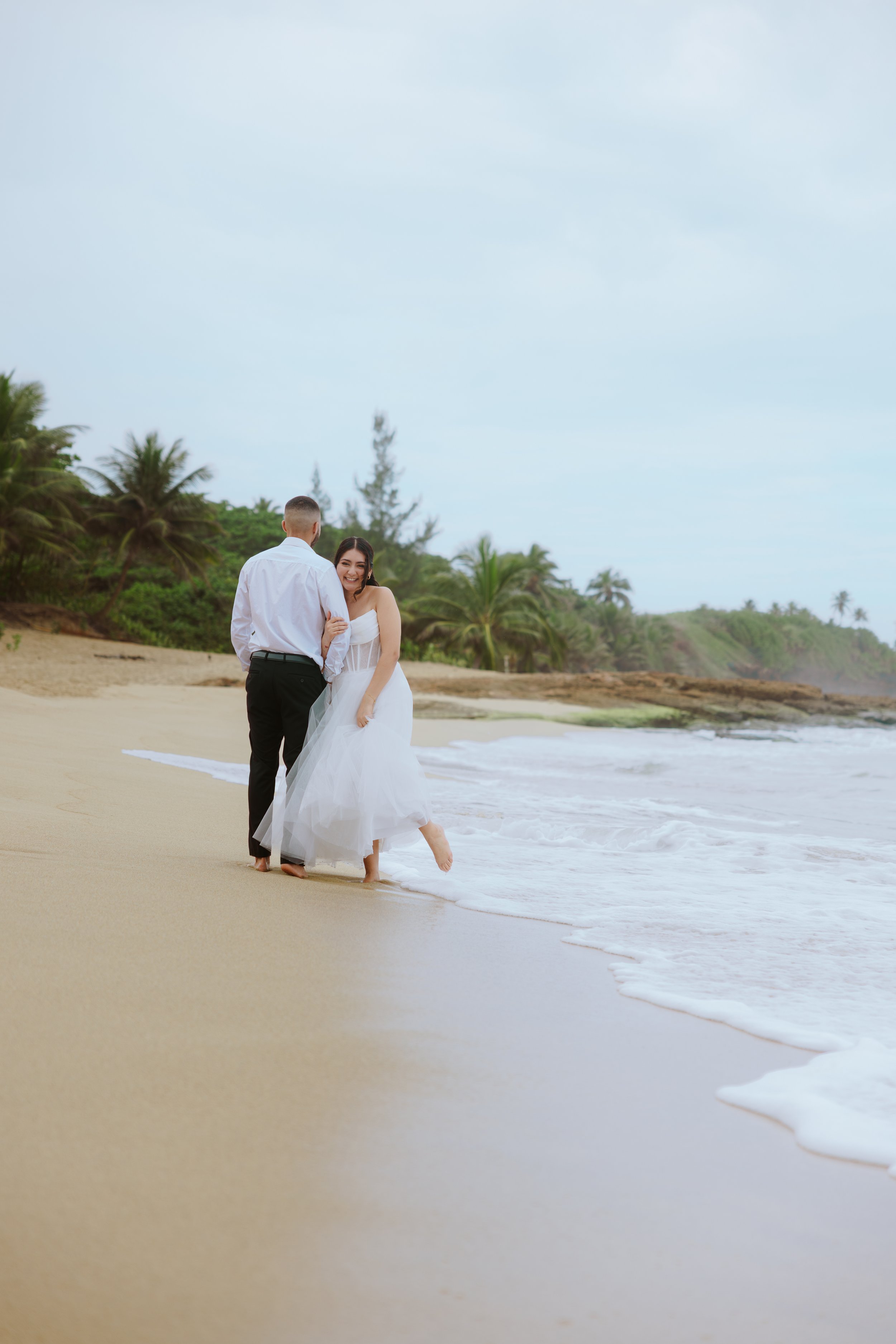 A couple dressed in wedding attire standing on a beach, smiling, with ocean waves at their feet, with lush green trees and a cloudy sky in the background.