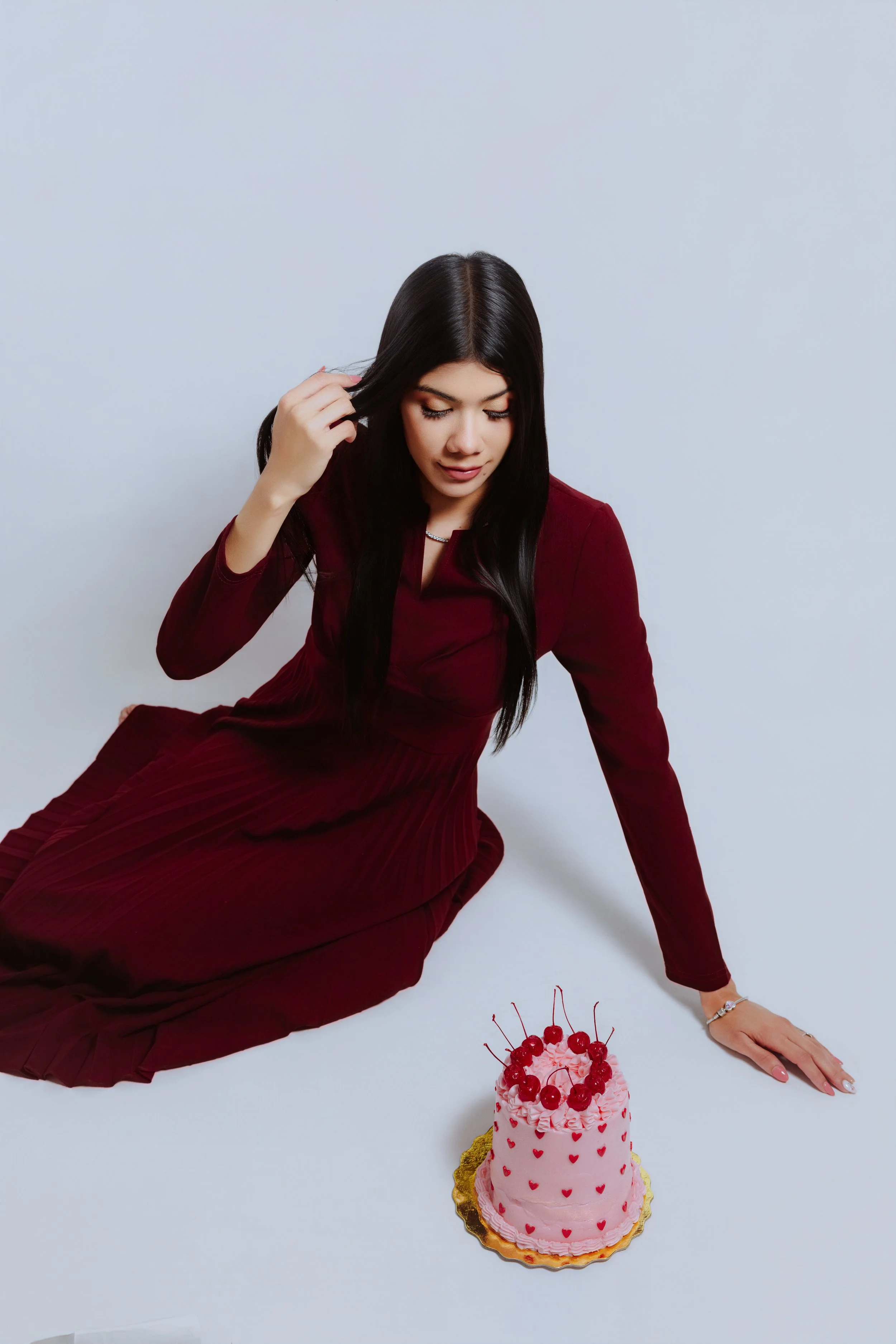 Young woman in a burgundy dress sitting on the floor with a pink heart-themed cake topped with cherries in front of her, against a plain white background.