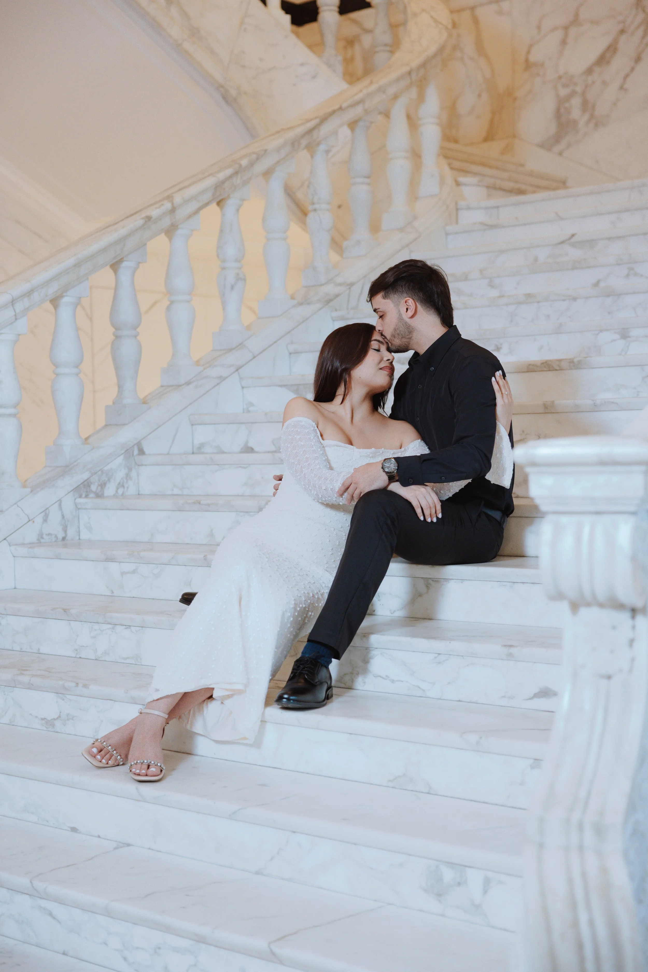 A romantic couple sitting on marble stairs, with the man kissing the woman's forehead. The woman is wearing a white off-shoulder dress and heels, while the man is dressed in black. The setting is elegant, with a marble staircase and ornate balustrade