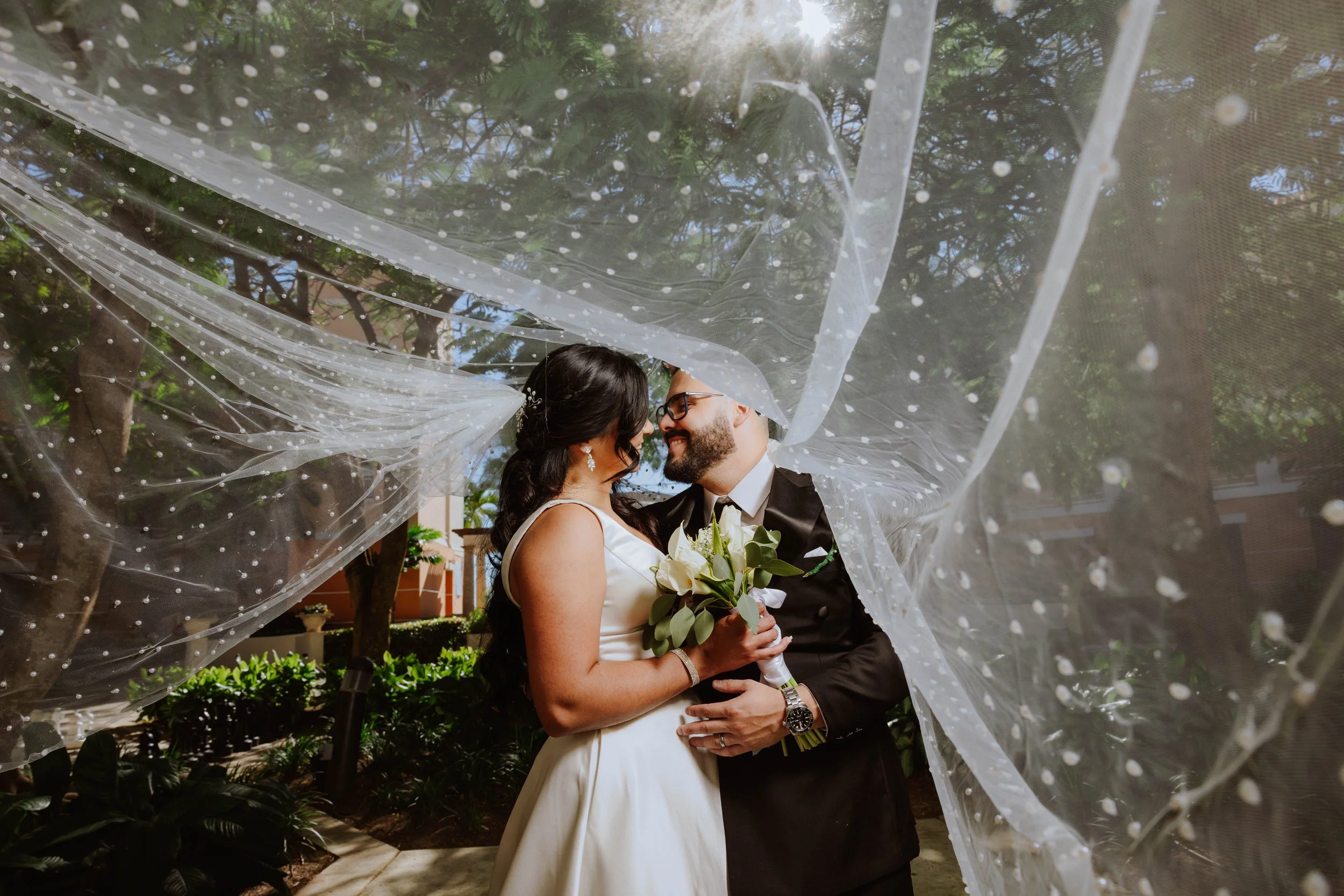 A bride and groom standing closely together under a flowing, dotted sheer fabric outdoors with trees in the background, sharing a tender moment.