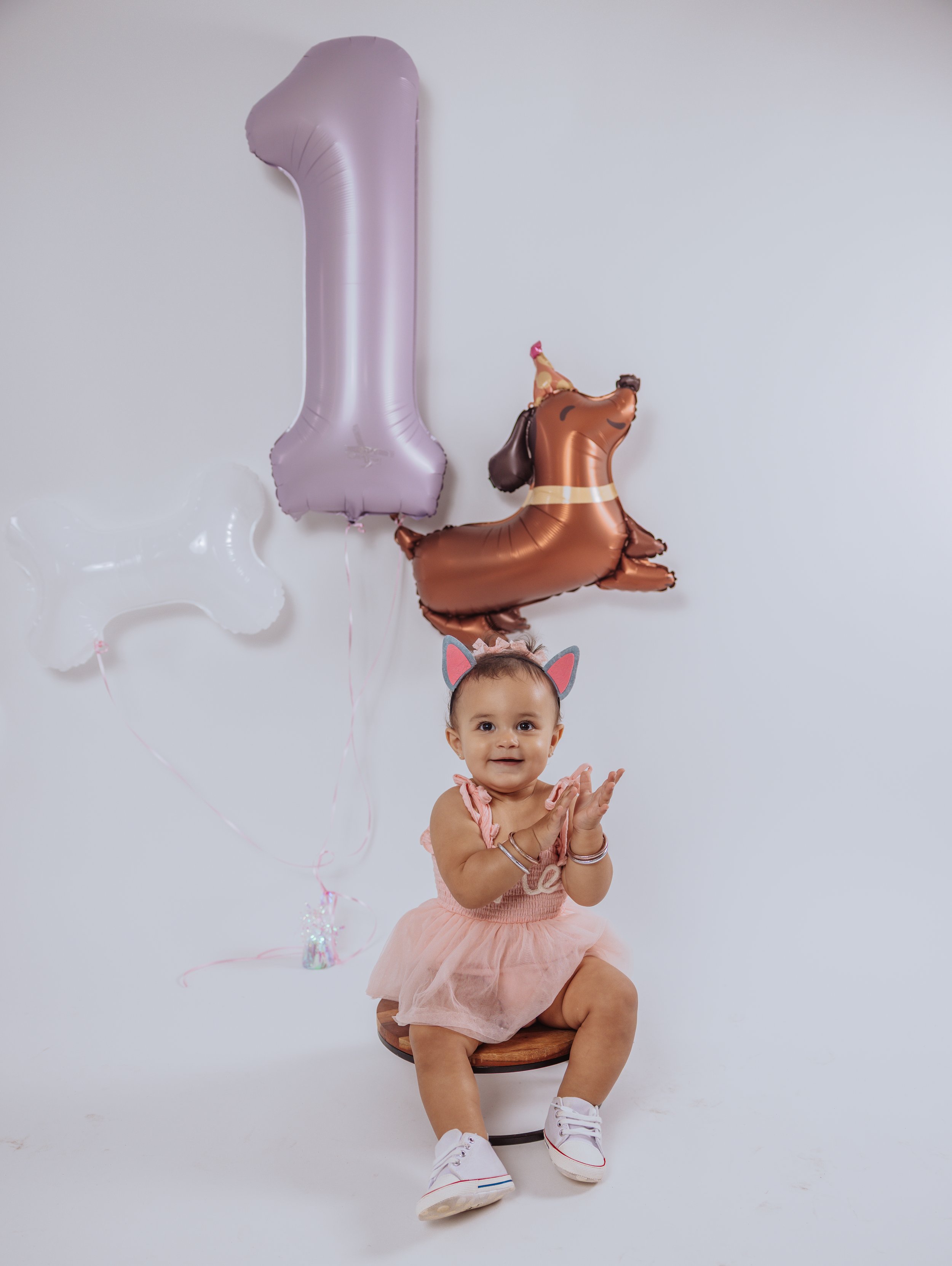 A young girl with cat ear headband sitting on a stool, wearing a pink dress, white sneakers, and bracelets, clapping her hands. Behind her are balloon animals of a horse, a giraffe, and a cloud, along with a large pink number one balloon.