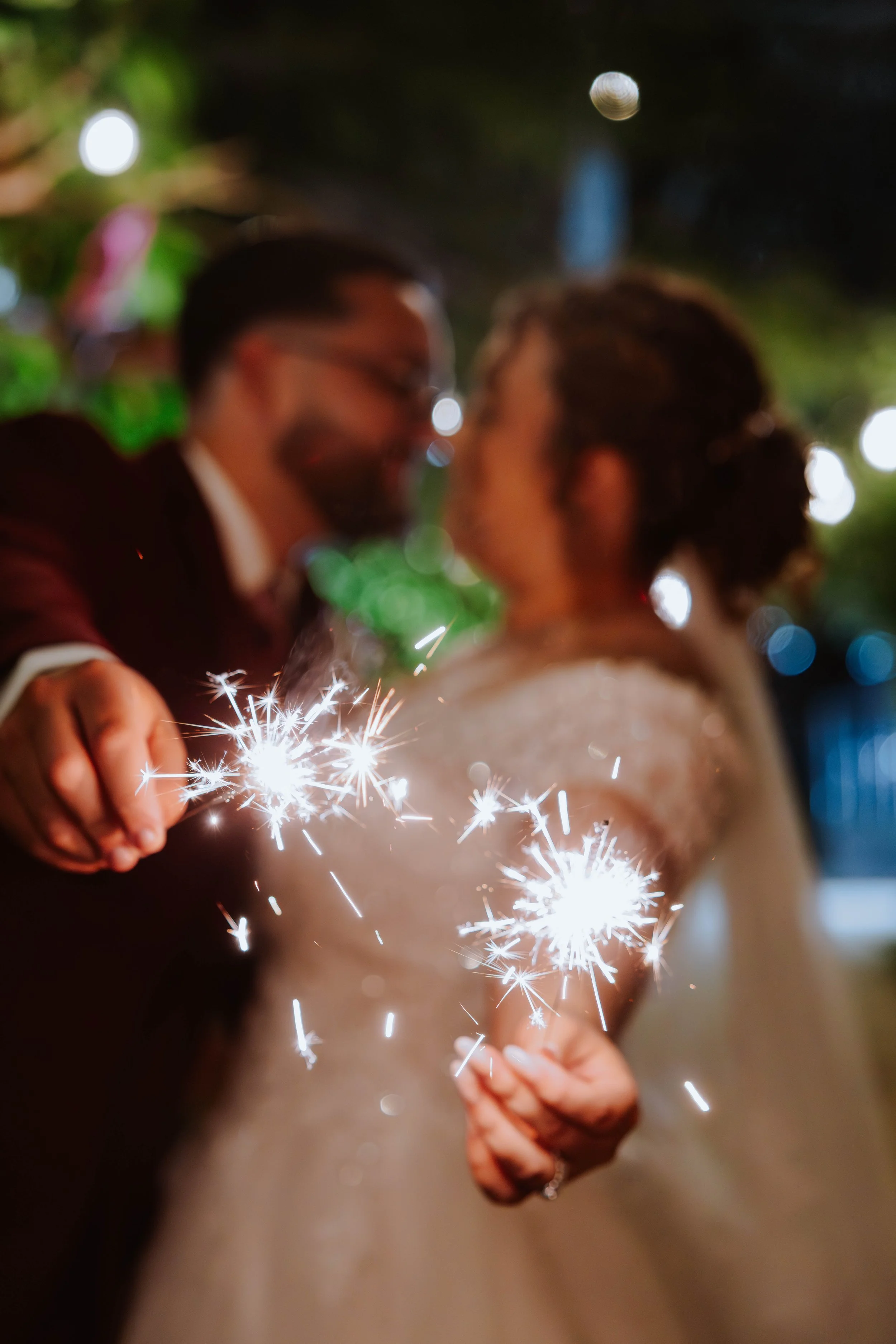A couple holding sparklers and smiling at each other at night, with blurred lights in the background.