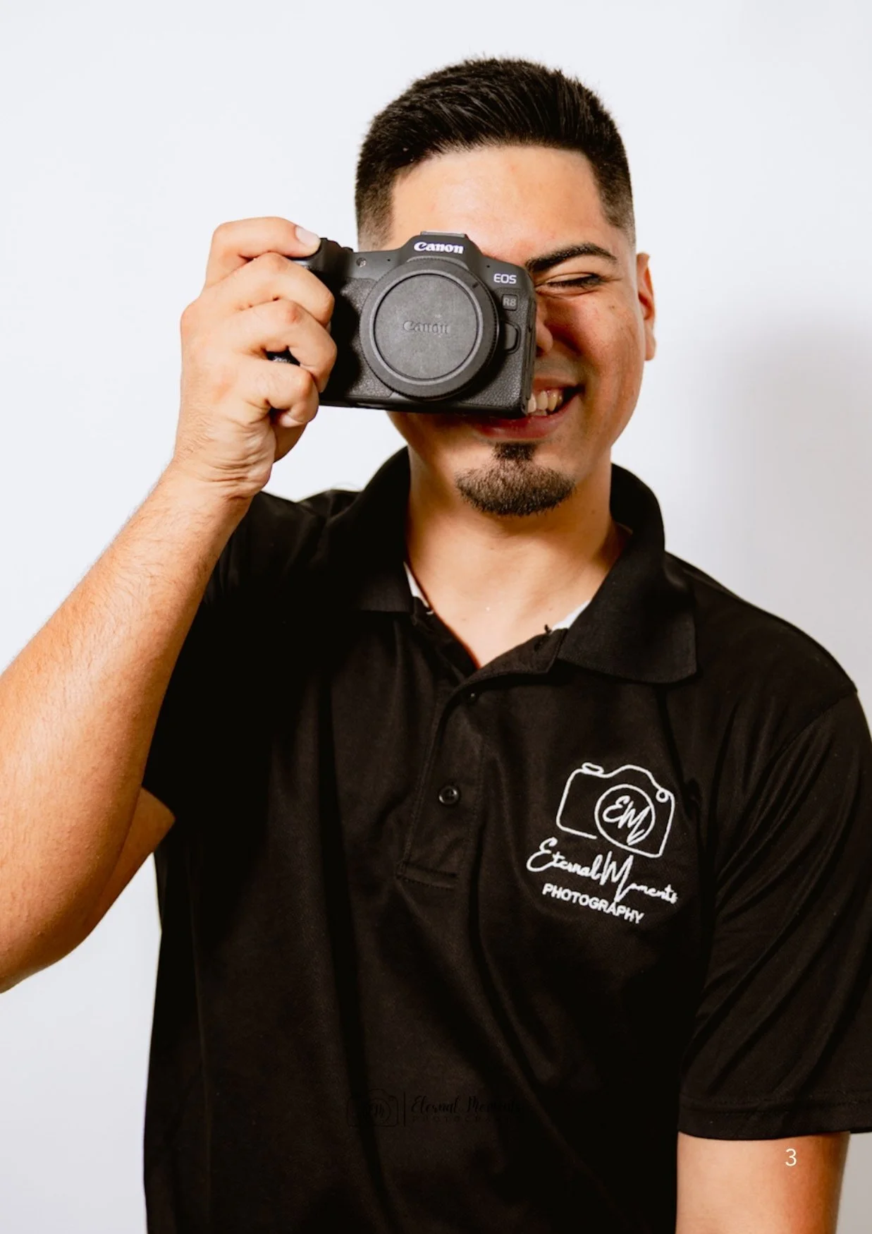 A smiling man holding a Canon camera up to his eye, with the camera lens covering one of his eyes, wearing a black shirt with a photography logo.
