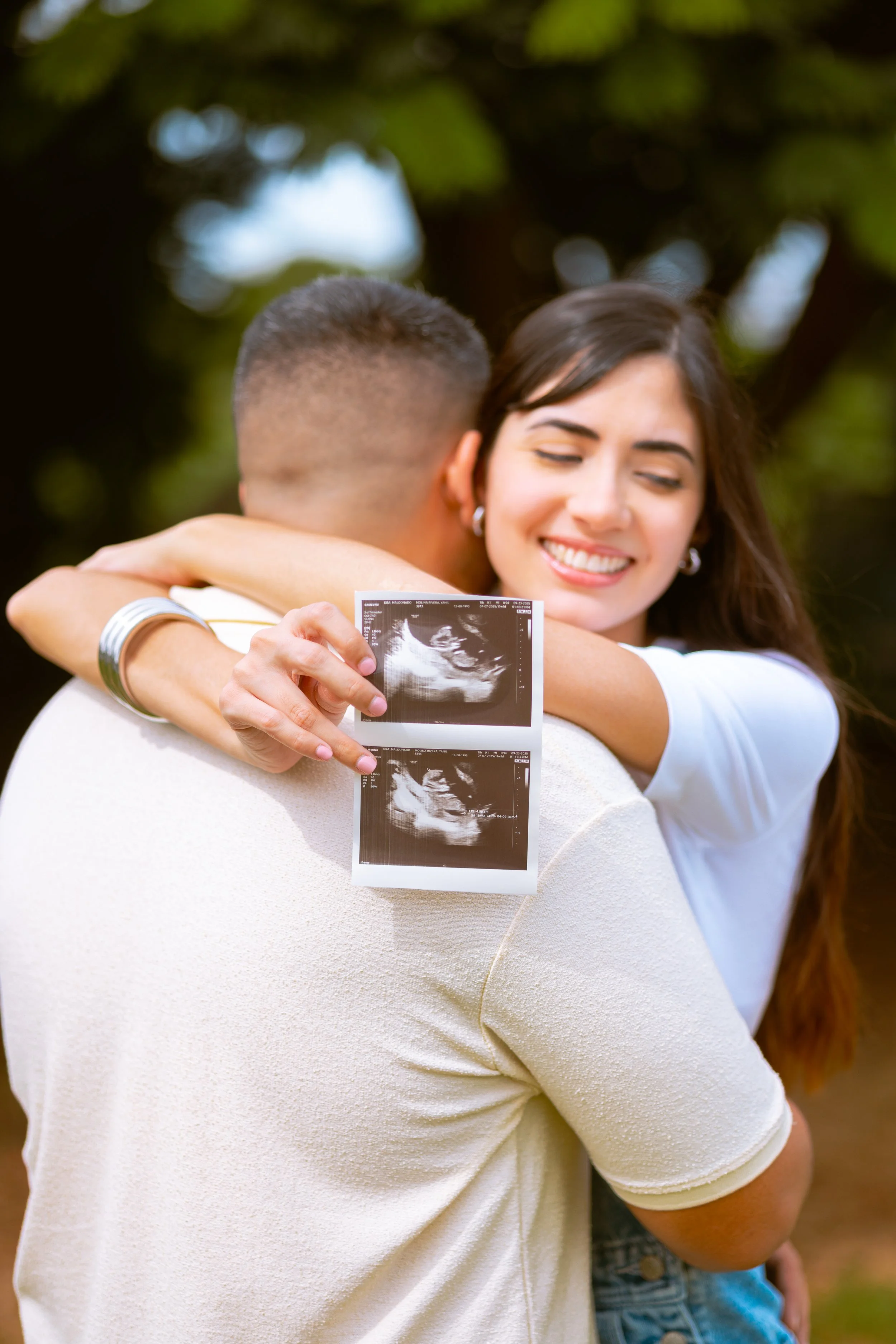 A woman embraces a man holding an ultrasound image in an outdoor setting with trees in the background.