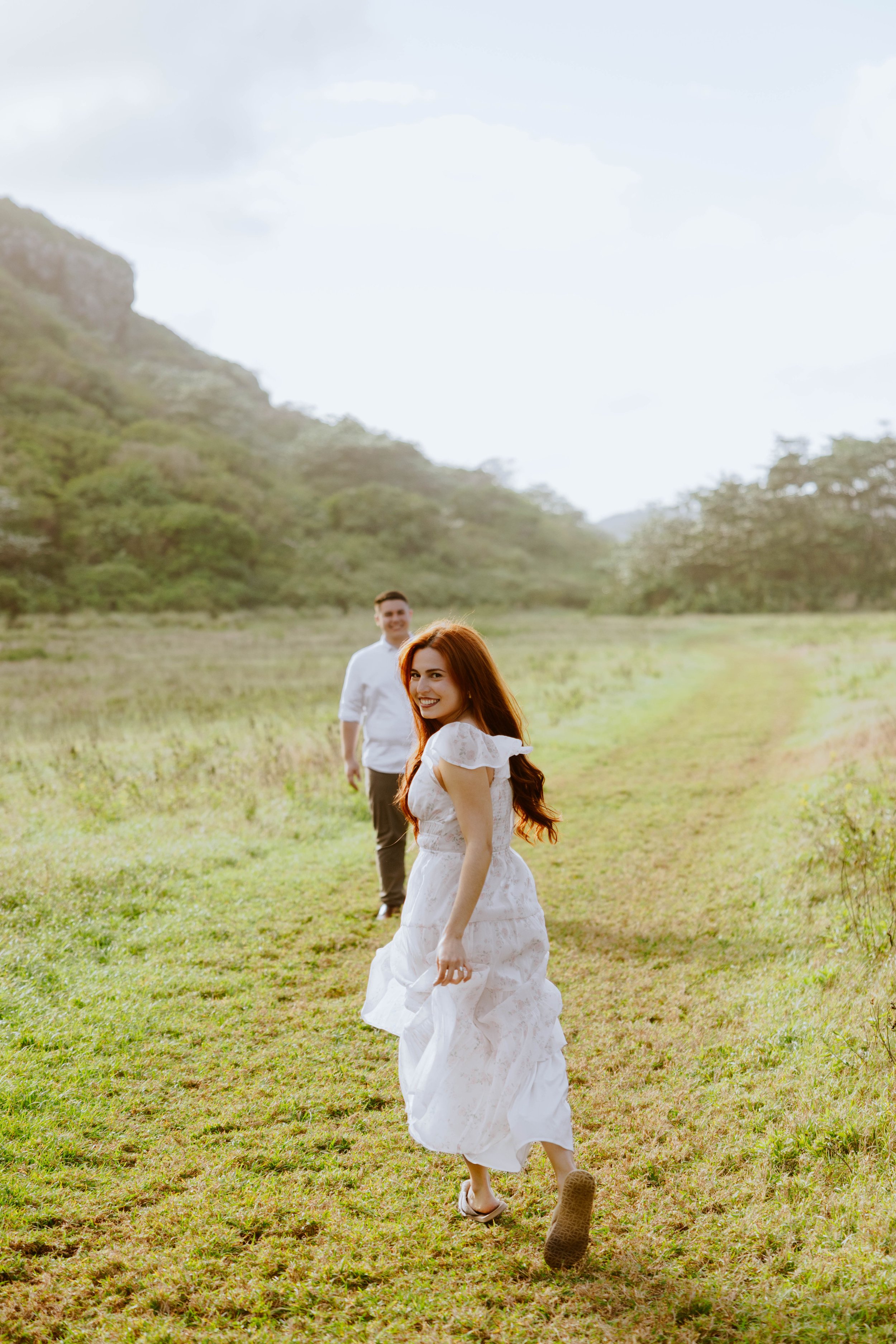 Young woman with red hair in a white dress smiling and walking through a green field with rolling hills and a cloudy sky in the background, with a man walking behind her.