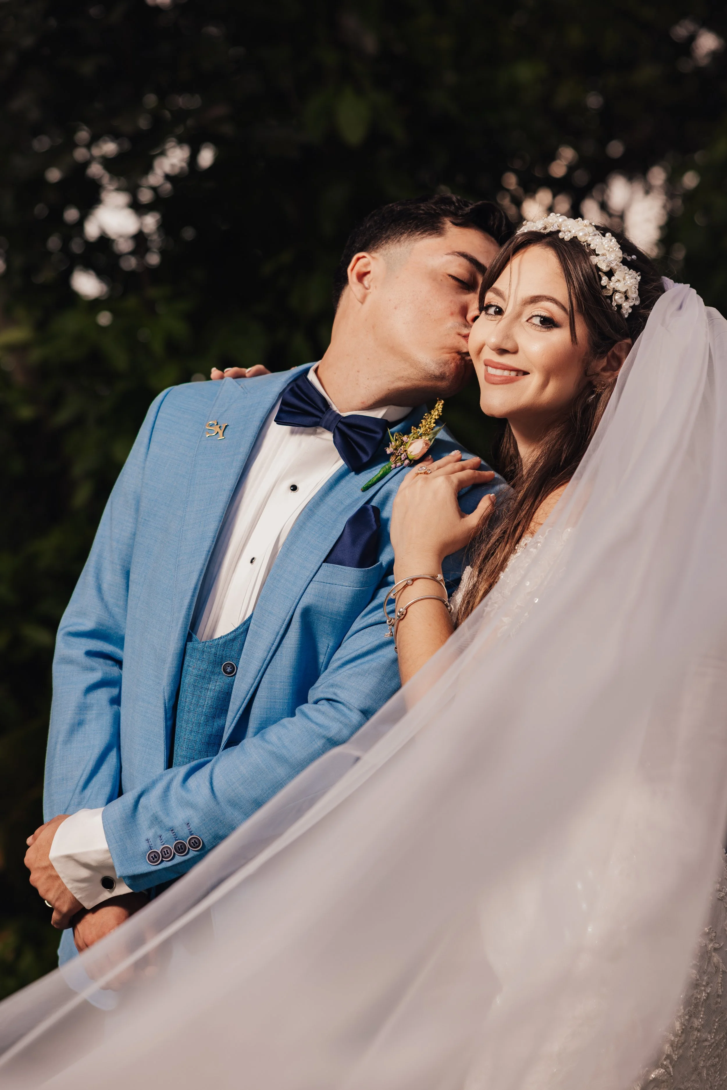 A bride and groom share a kiss outdoors, with the groom dressed in a blue tuxedo and the bride in a white wedding gown with a floral headpiece. They appear to be happy and celebrating their wedding.