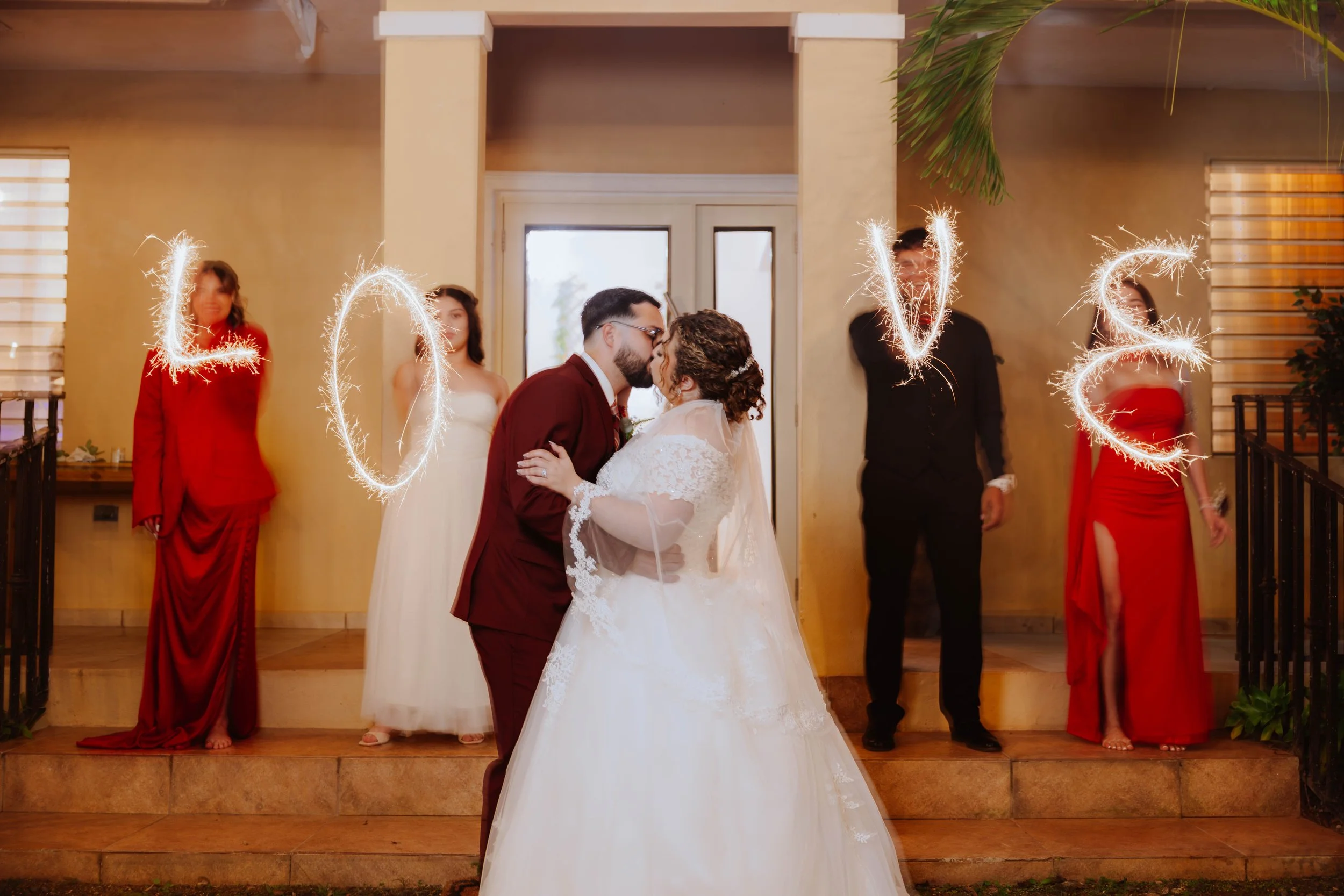 A bride and groom kiss at their wedding reception, with sparklers spelling 'LOVE' in the background, and four bridesmaids standing behind them in red dresses and one woman in a white dress.