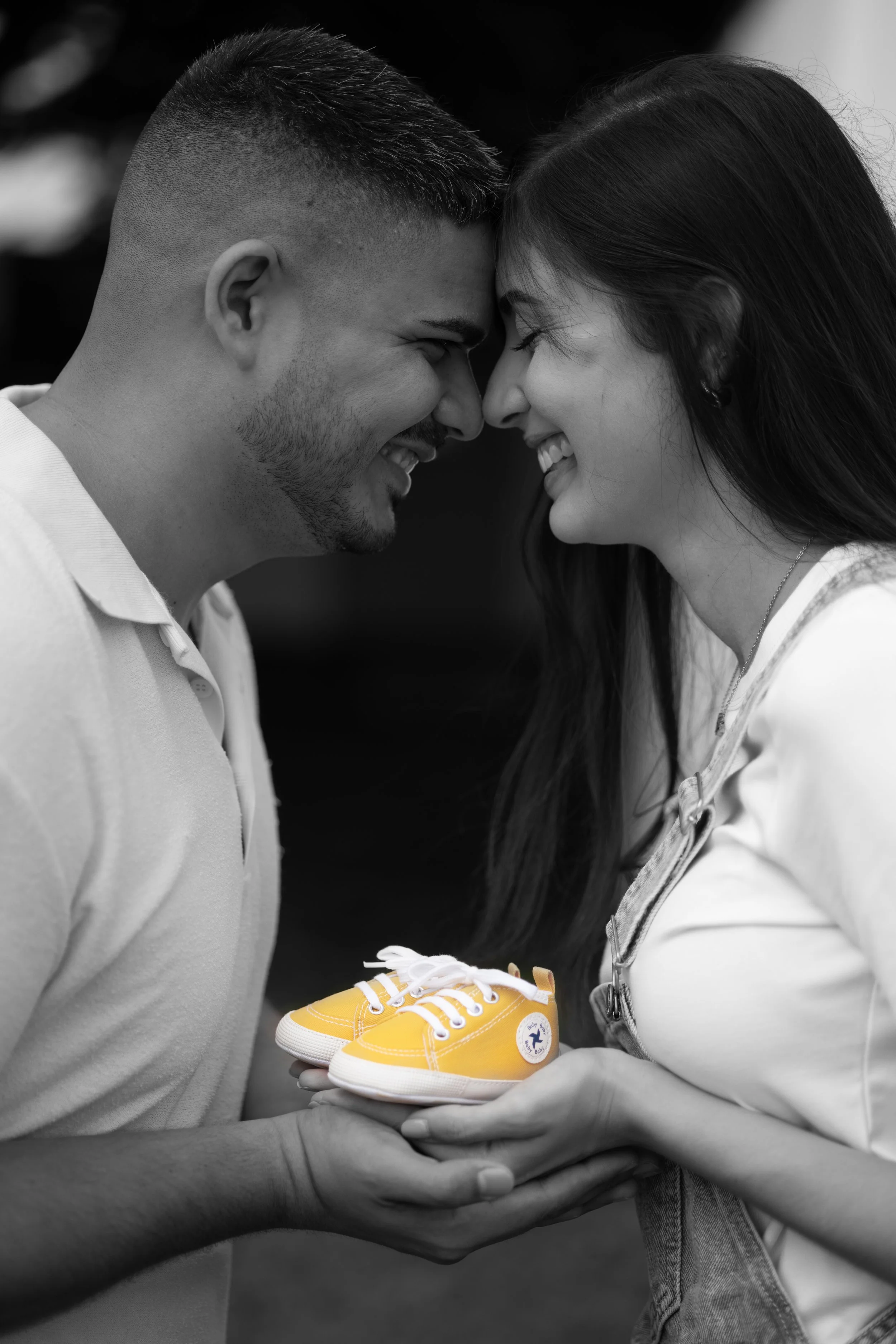 Close-up of a man and woman pressing foreheads together, smiling, with a pair of yellow toddler shoes being held between them.